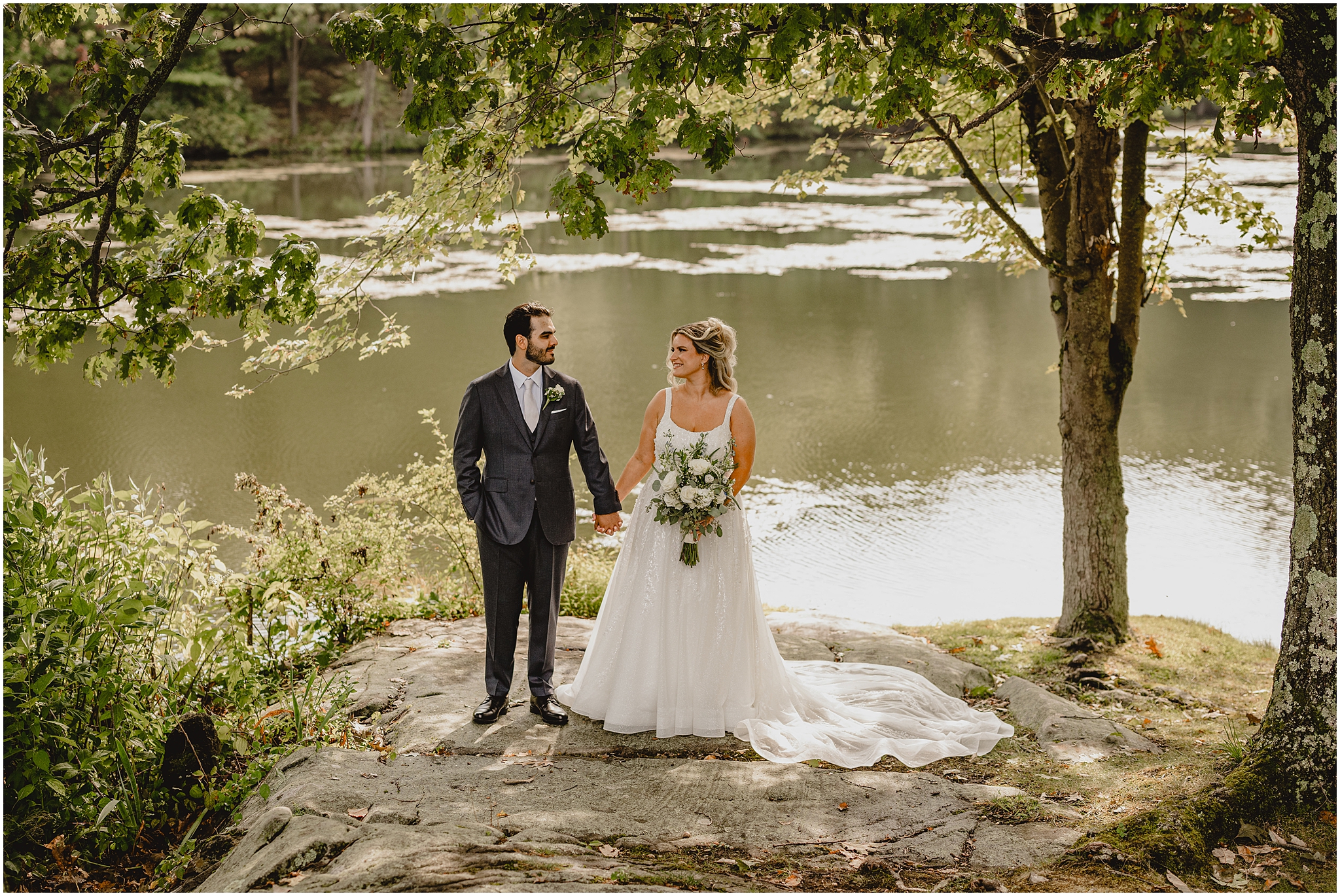 bride and groom hold hands standing on flat rock by pond at the Refinery at Perona Farms