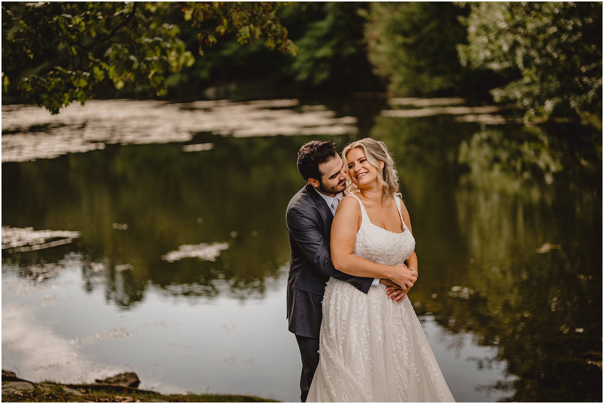 groom hugs bride from behind near pond at the Refinery at Perona Farms