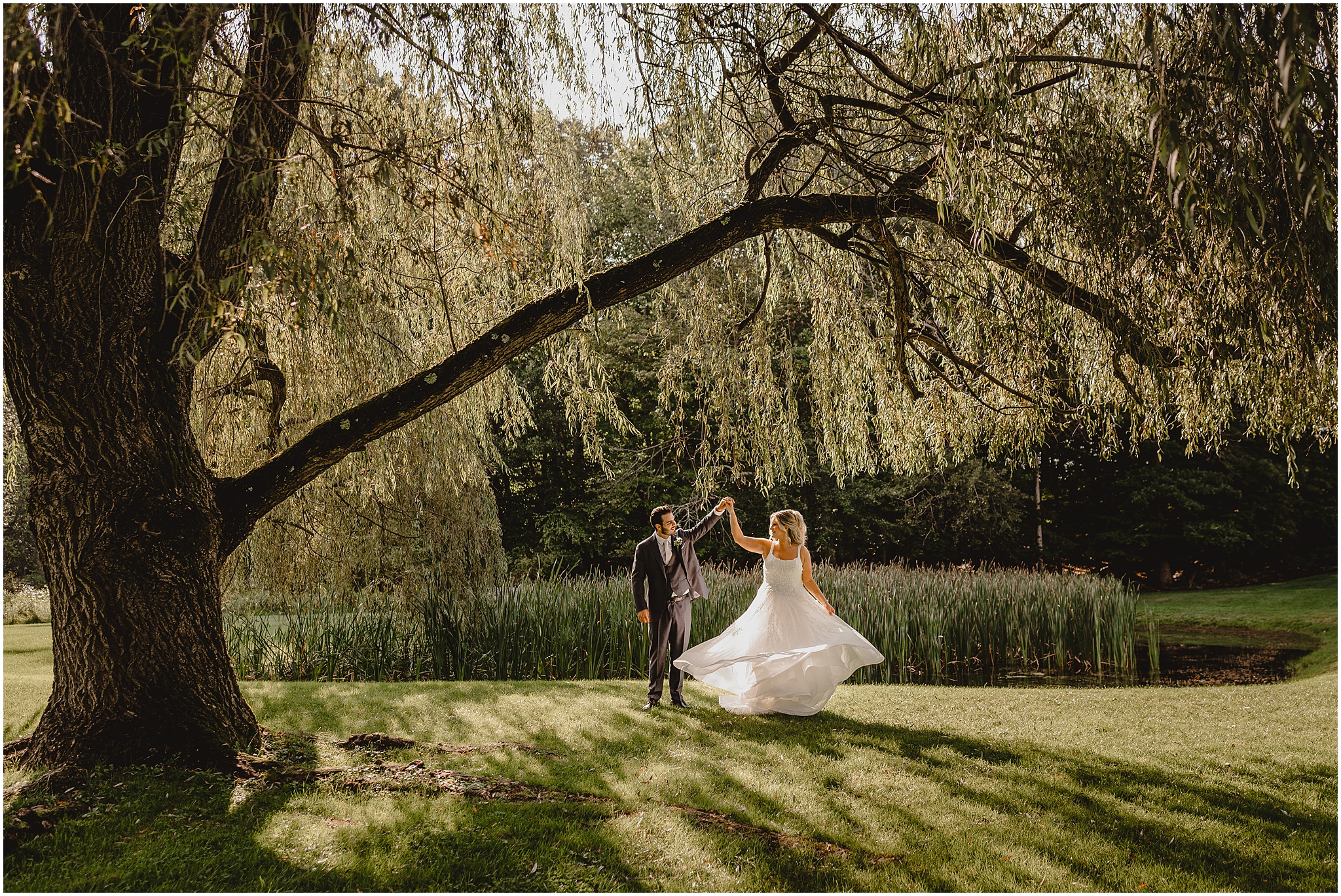 groom twirls bride under weeping willow by pond at the Refinery at Perona Farms