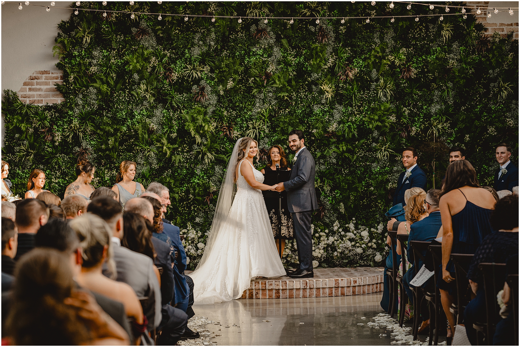 bride and groom hold hands during wedding ceremony on brick dais looking at guests 