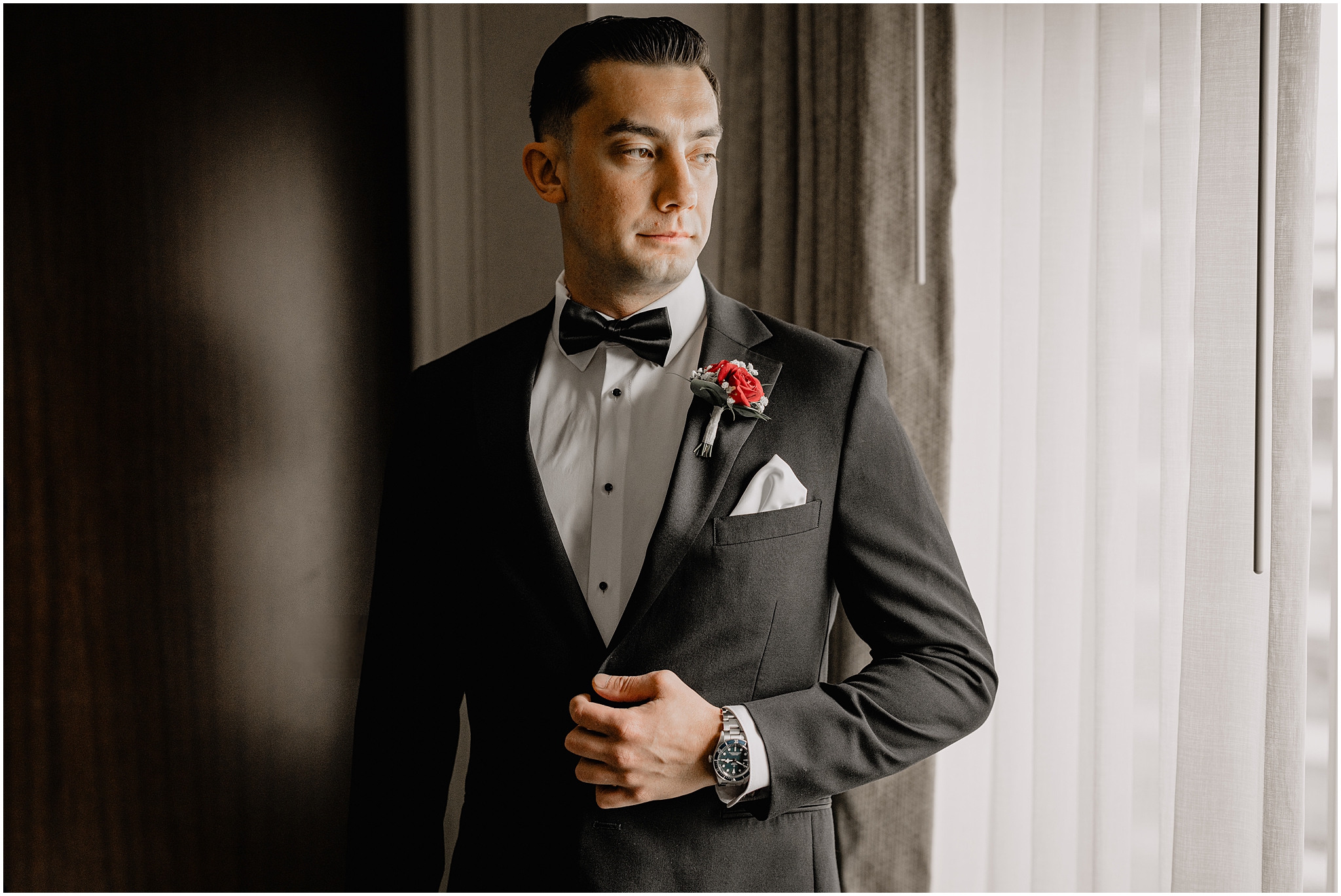 groom poses in black suit with red rose boutineere in front of hotel room window