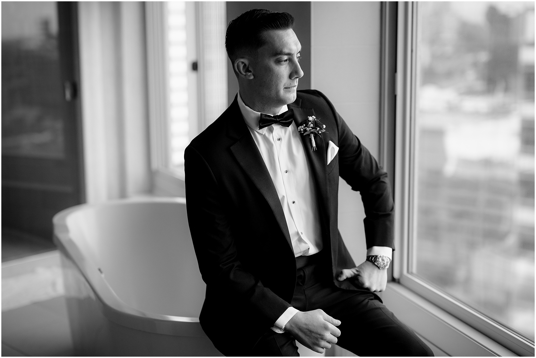 groom sits in front of hotel window on edge of bathtub in black tux