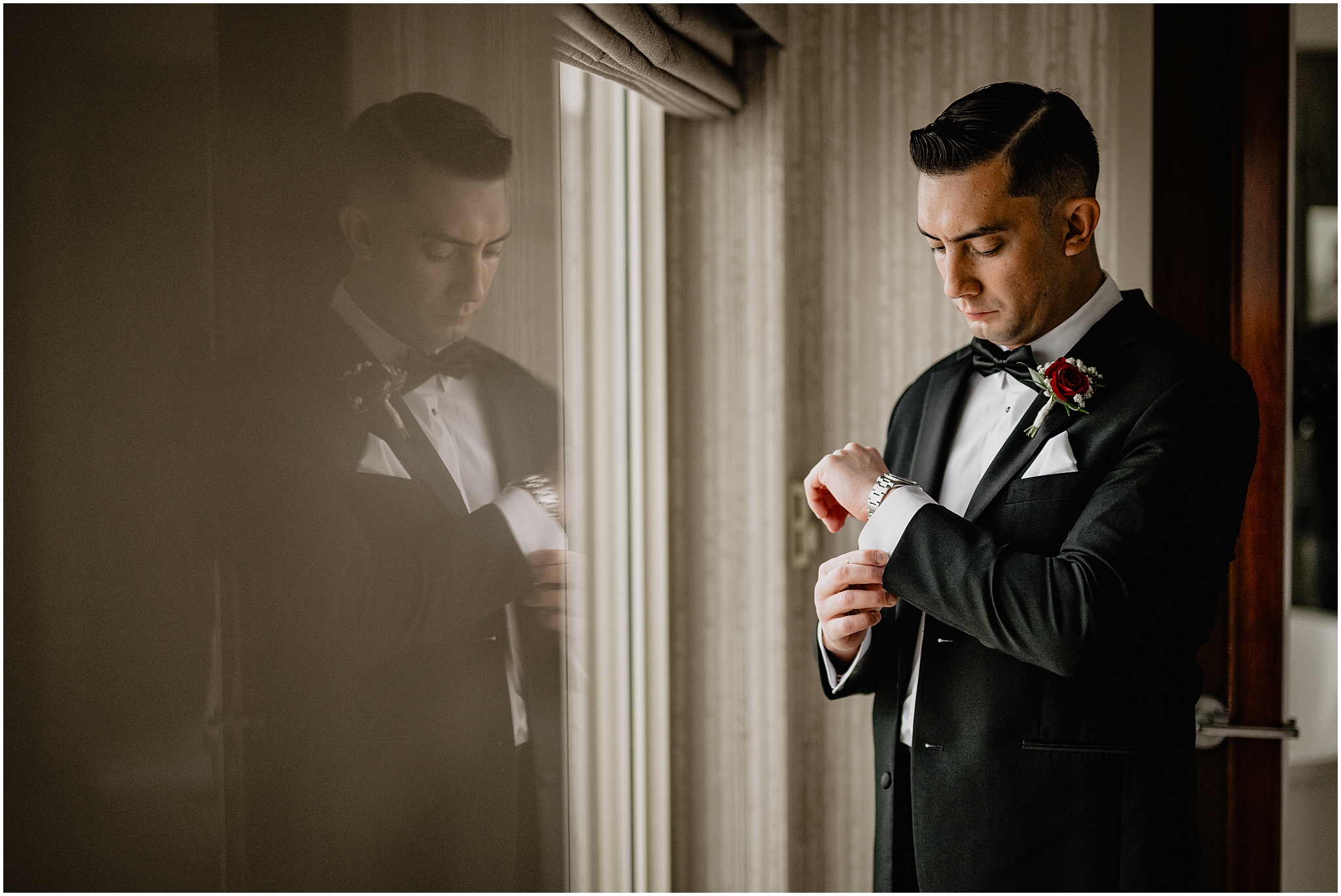 groom adjusts cufflinks in hotel room with reflection of him on window pane