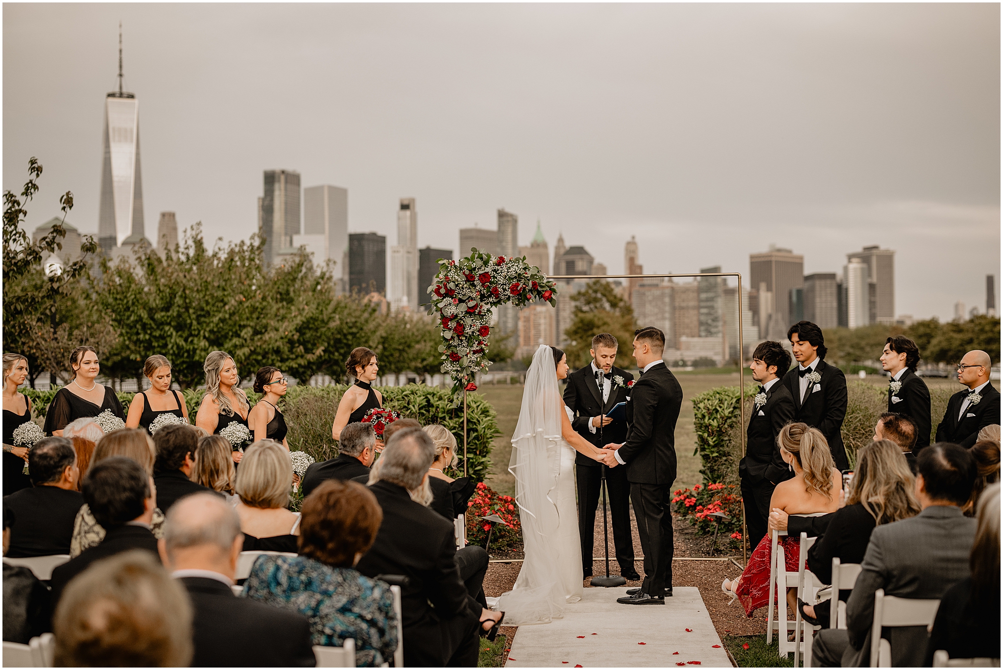 bride and groom hold hands in front of gold arbor with spray of red and white flowers on the corner at the Liberty House