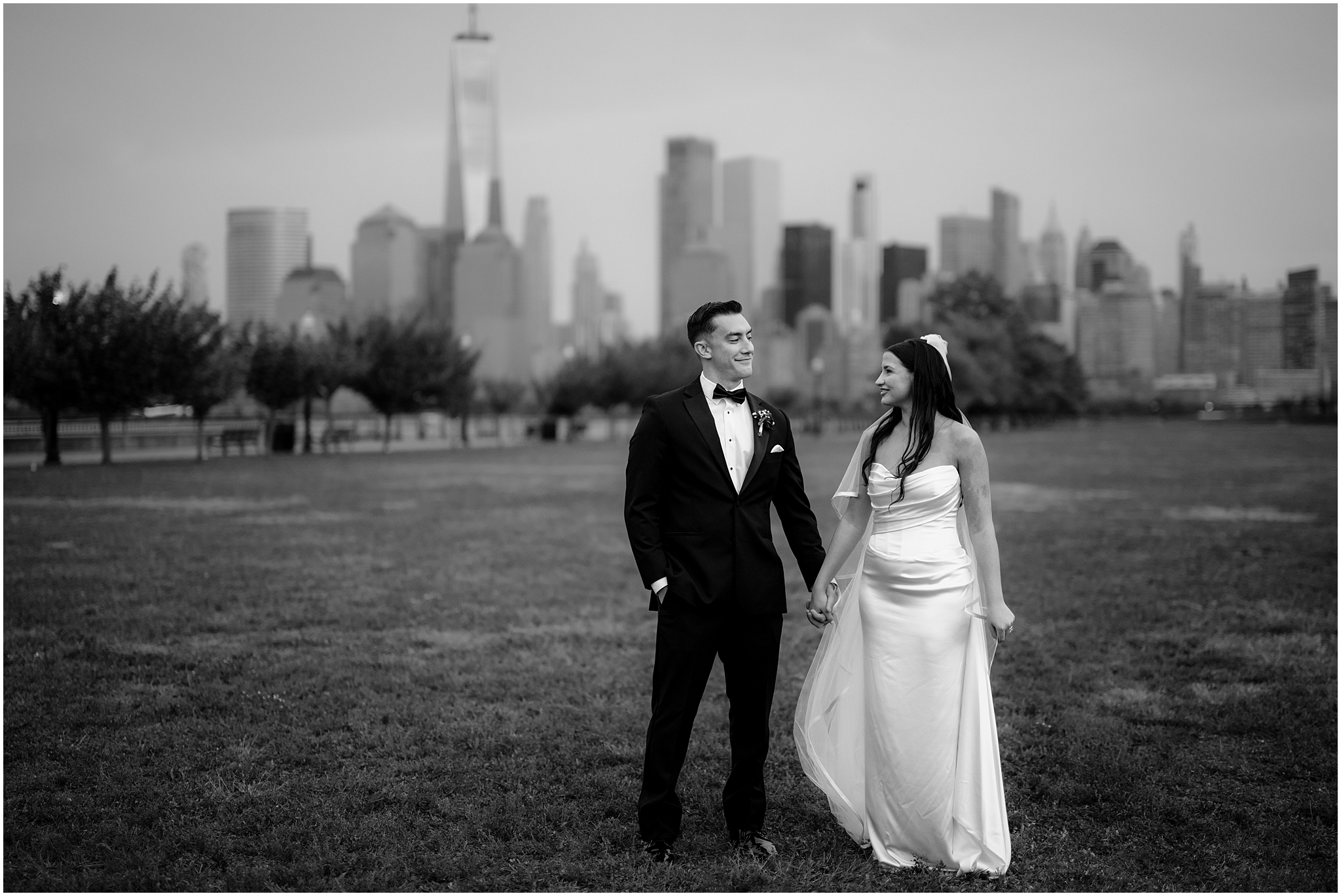 newlyweds hold hands looking at each other on lawn with Manhattan skyline behind them