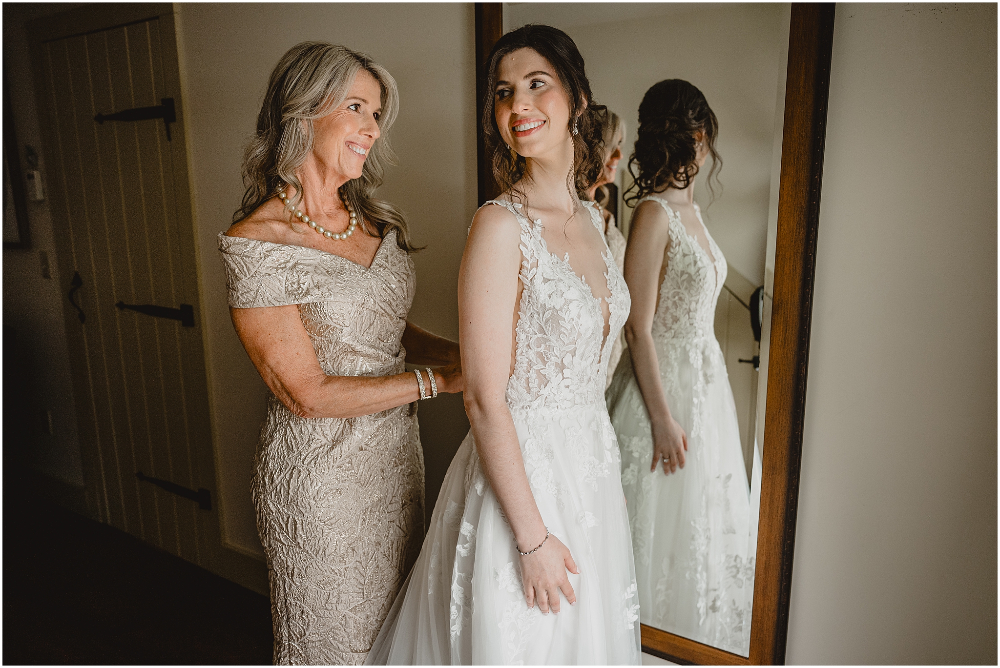 bride smiles over her shoulder at mom buttoning up her dress