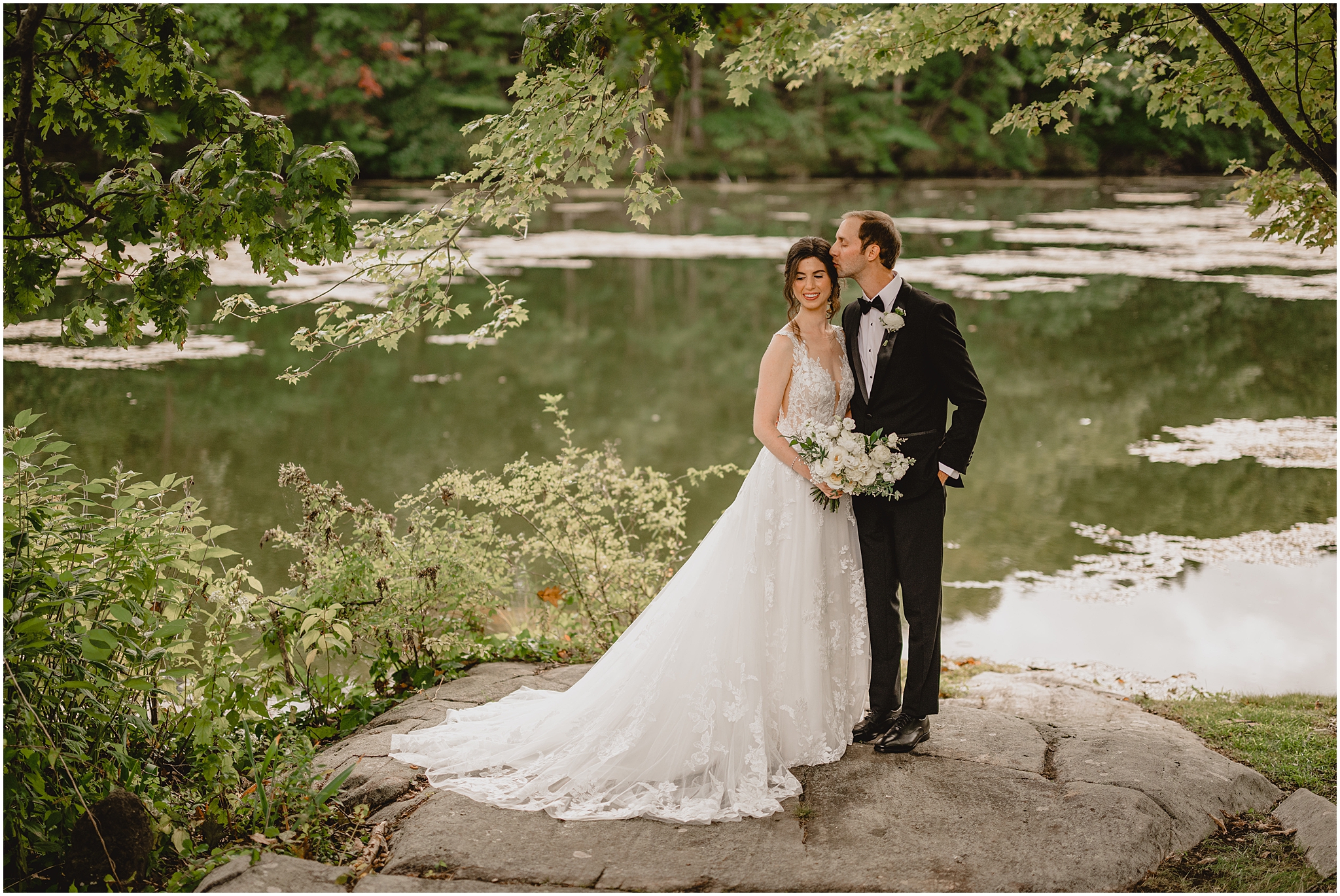newlyweds stand on flat stone while man kisses woman's head