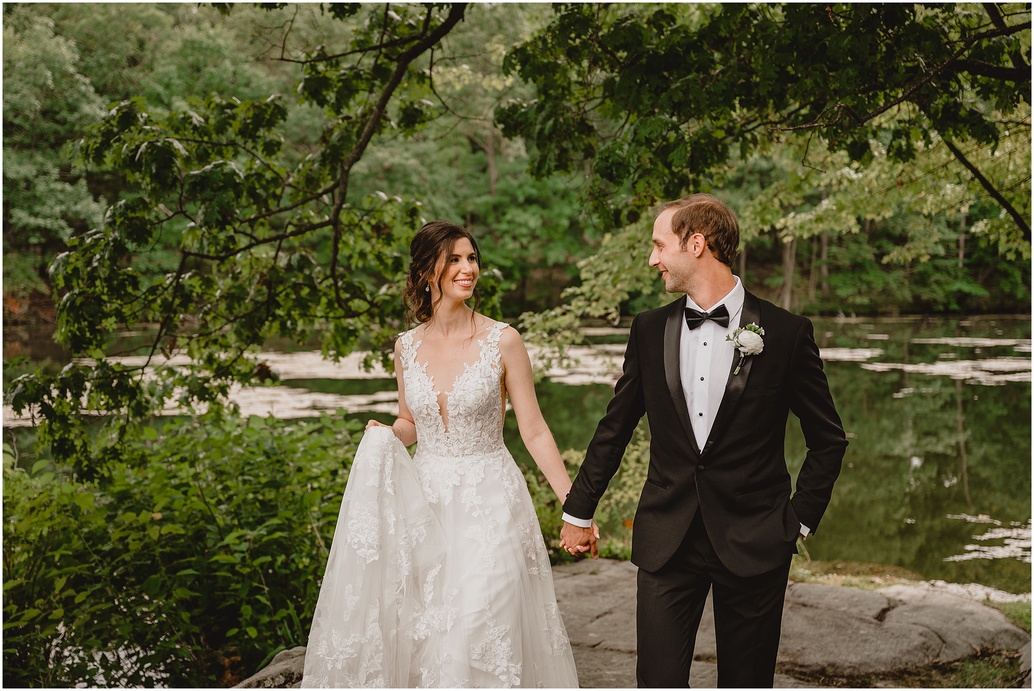 bride and groom hold hands standing on edge of pond