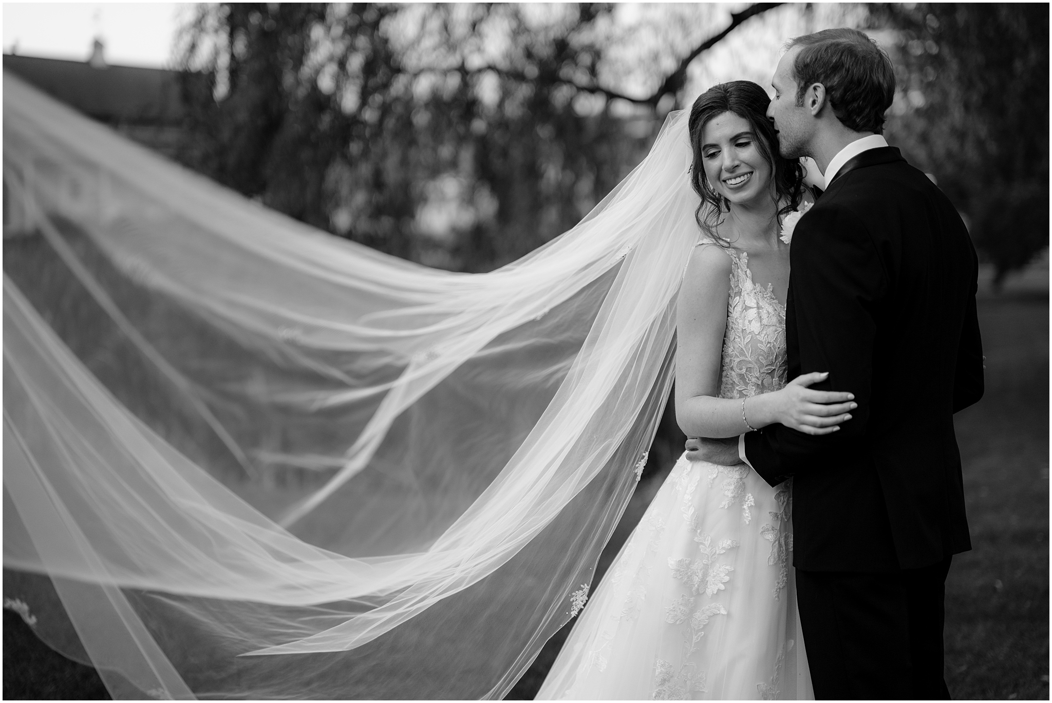 groom kisses bride's head while veil floats behind her on grounds of the Reserve at Perona Farms