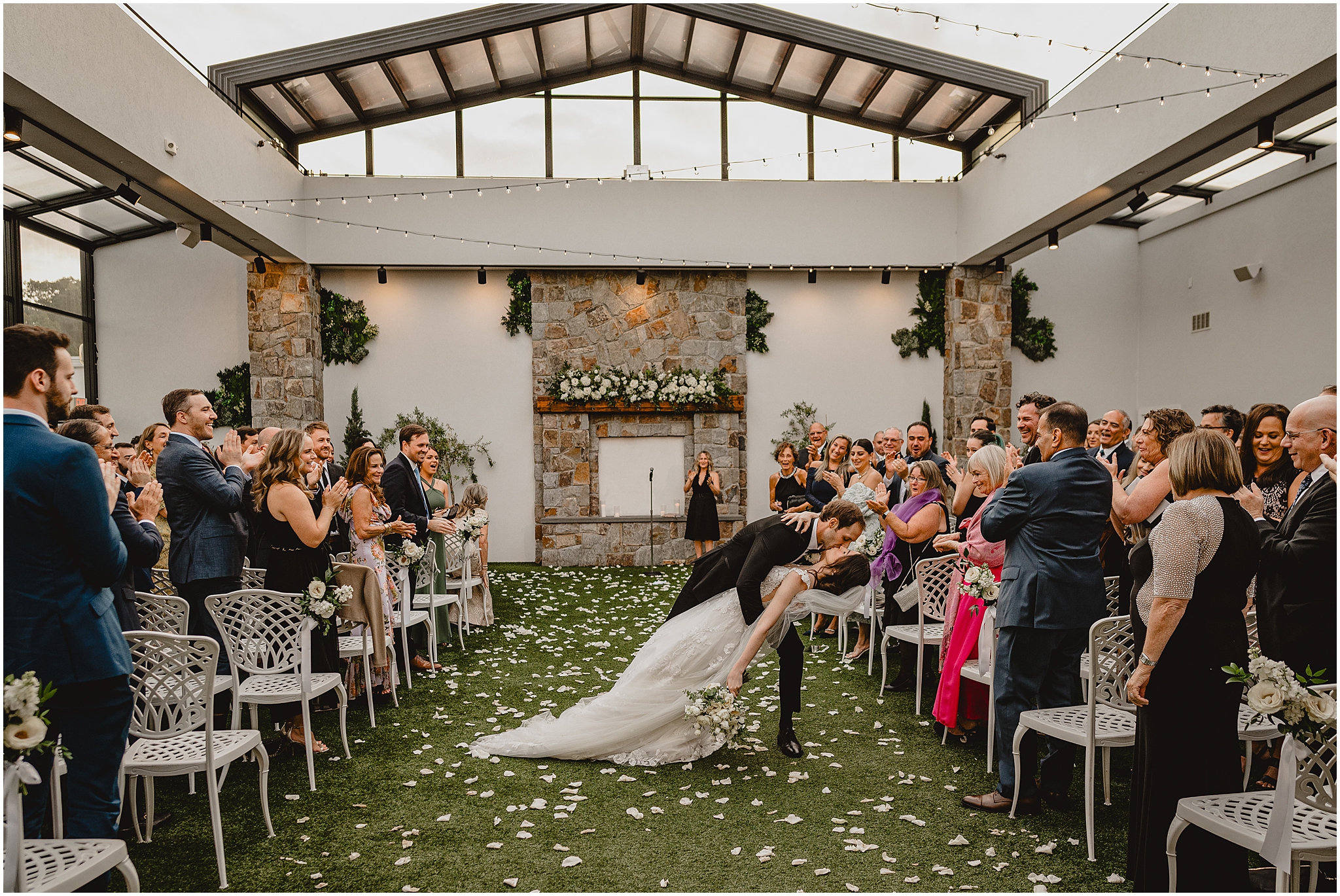 groom dips bride in aisle covered with white flower petals in atrium at the Reserve at Perona Farms