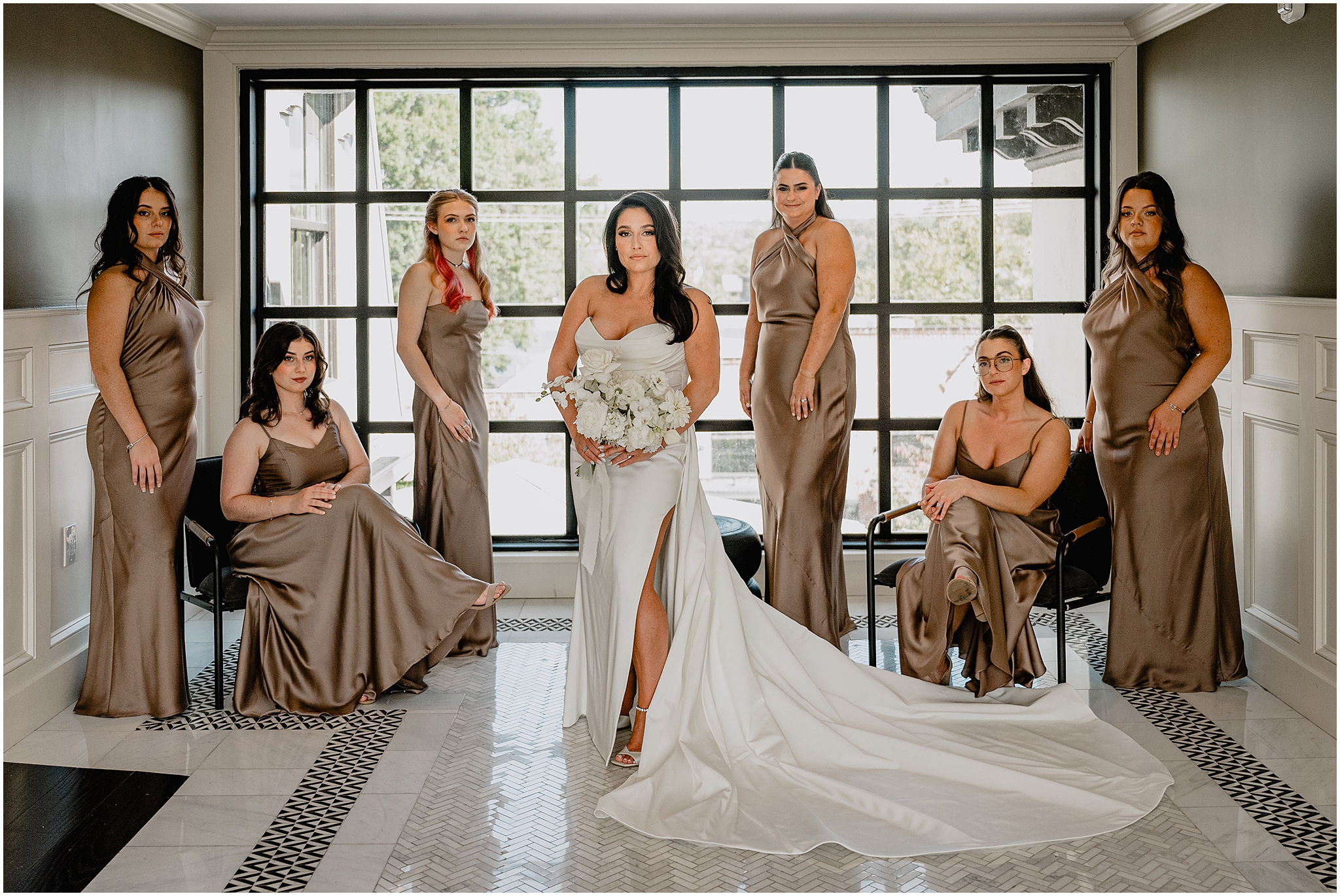 bride stands in front of bridesmaids in mismatched satin mocha gowns in front of window at The Logan Inn
