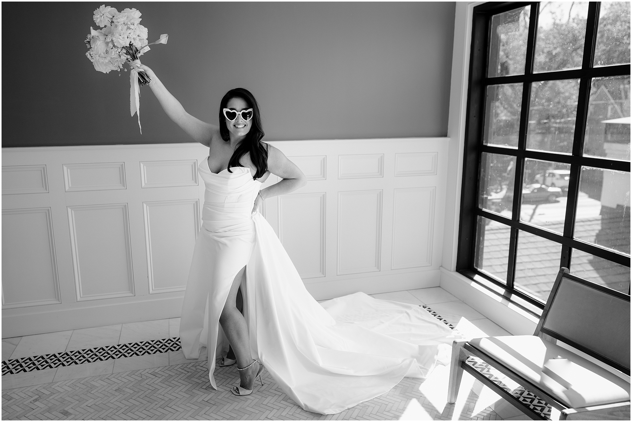 bride grins holding bouquet of white flowers in the air wearing heart shaped sunglasses in strapless gown 