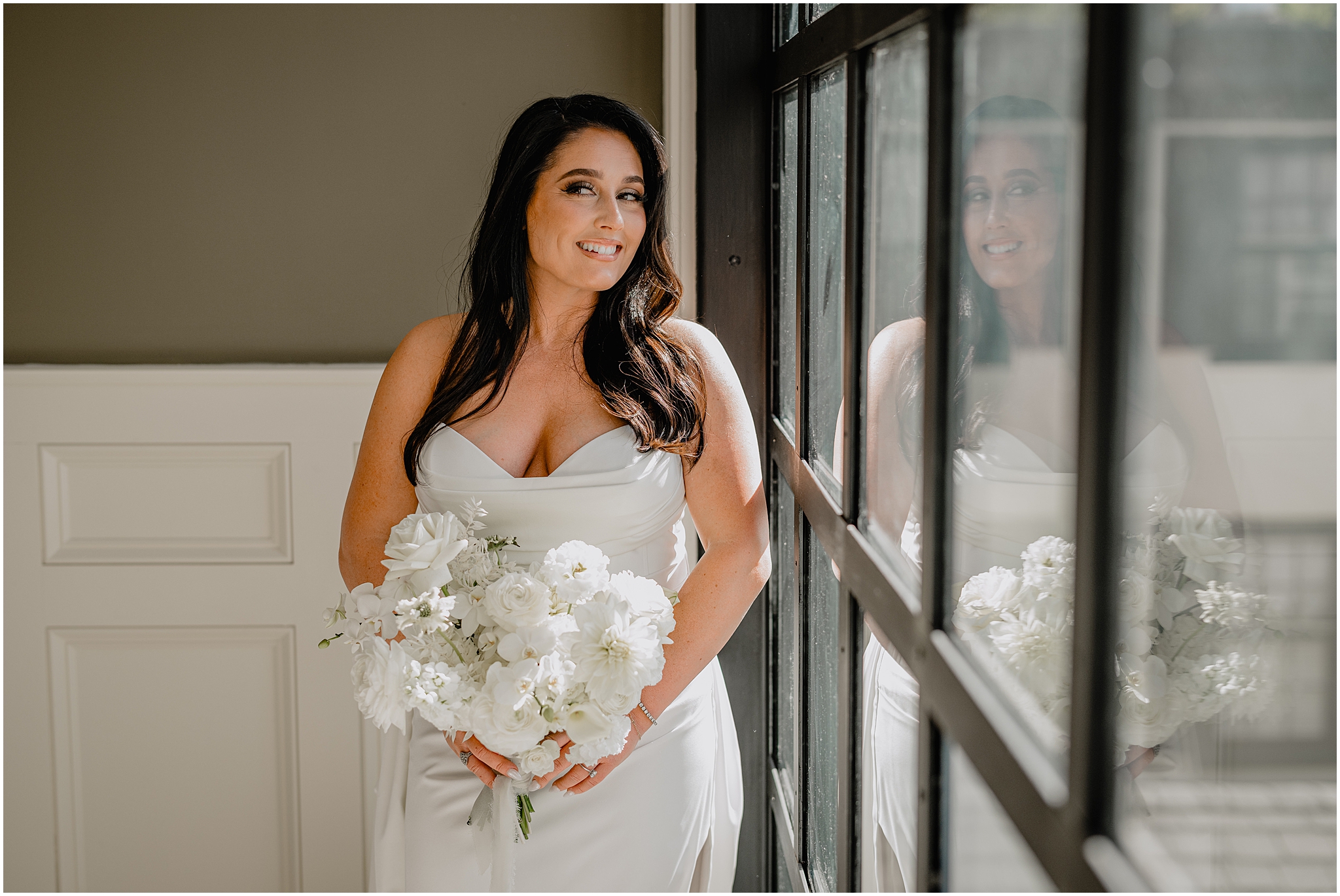 bride holds bouquet of all white flowers in stylish strapless gown next to window with black panes