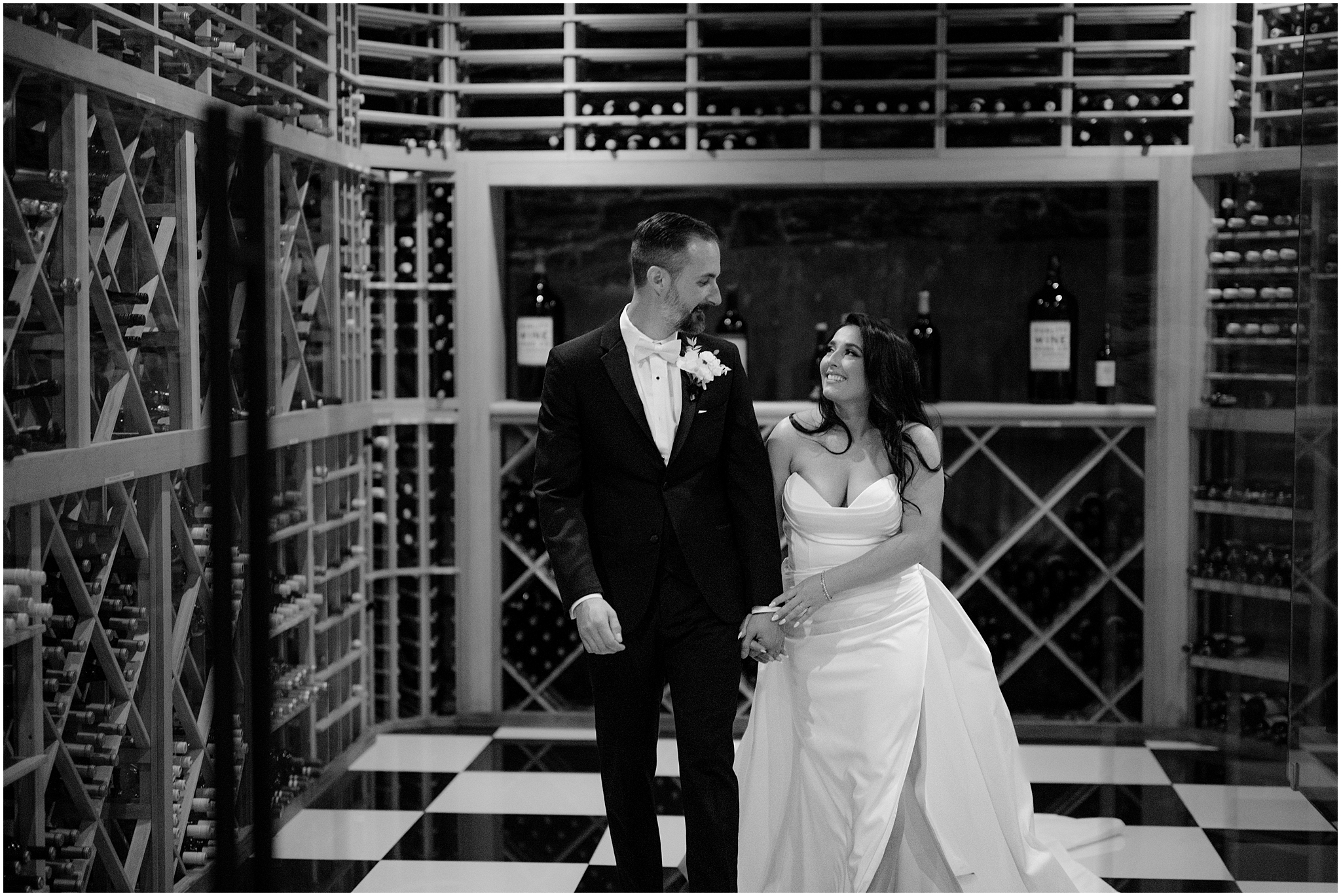 bride and groom hold hands walking in front of displays of wine bottles in wine cellar 