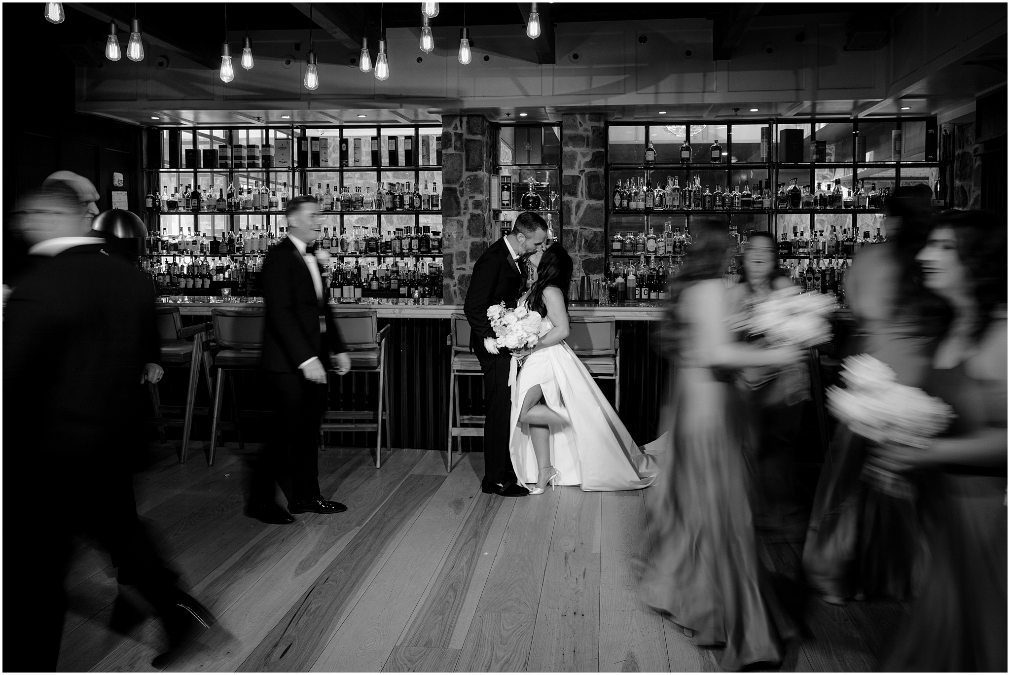 newlyweds kiss while wedding party walks around them blurred in black and white photo