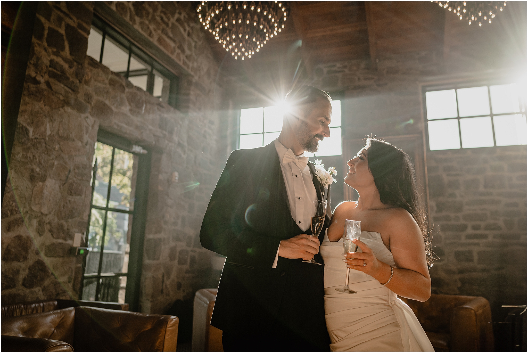 newlyweds hold glasses of champagne while smiling at each other in stone room at The Logan Inn