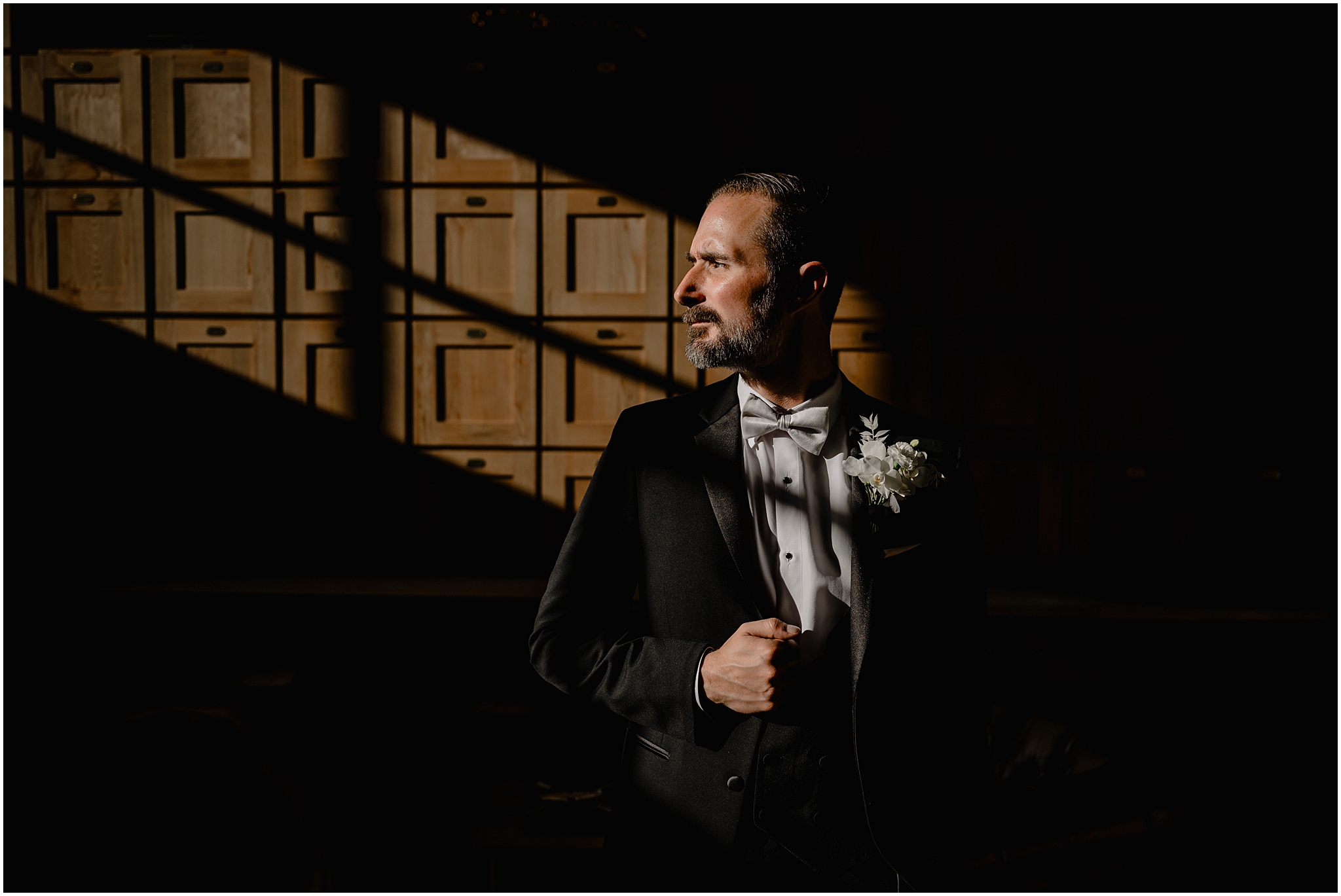 groom stands in front of wood paneling holding lapel of black suit 