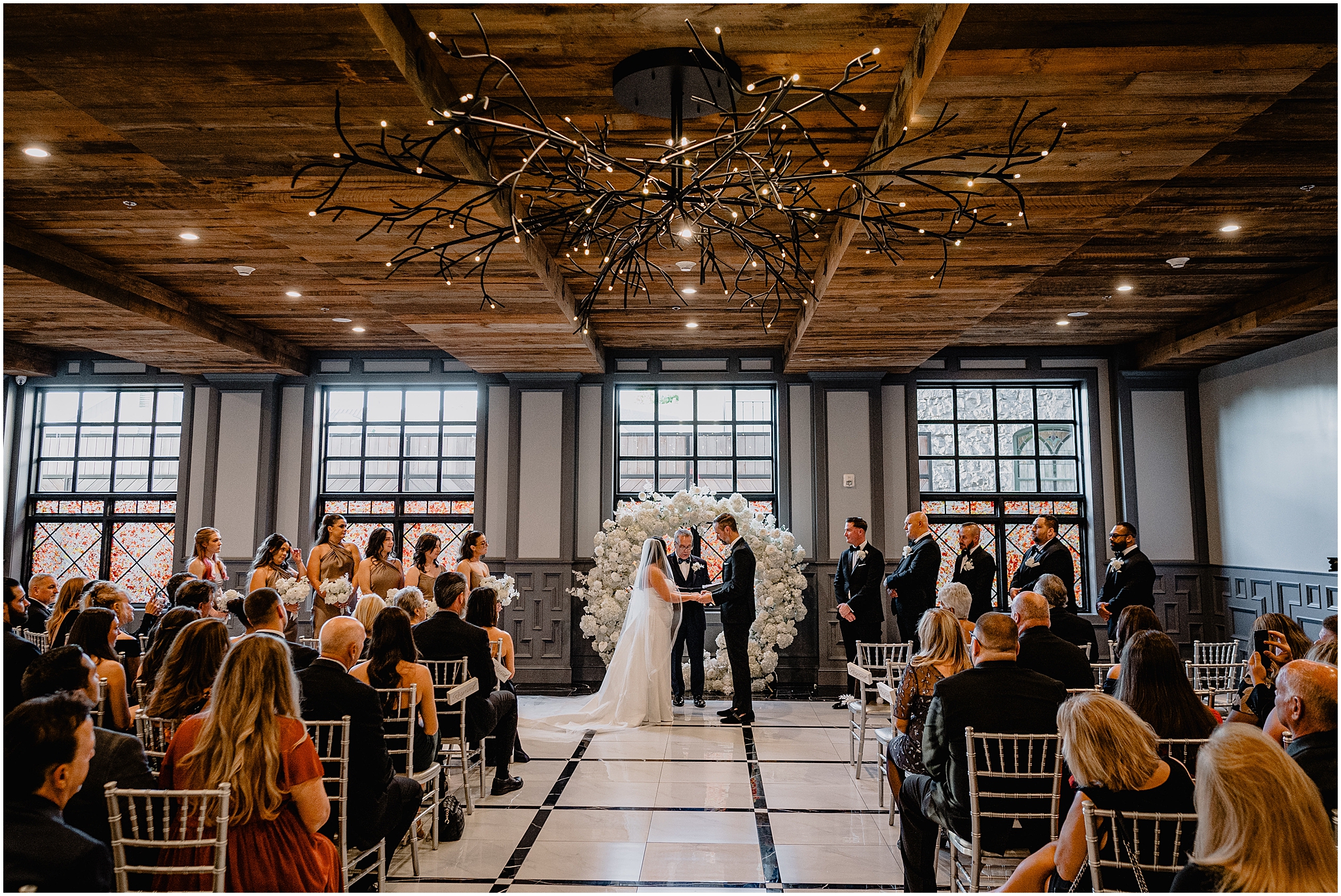 bride and groom hold hands exchanging vows during wedding ceremony in front of white floral arbor