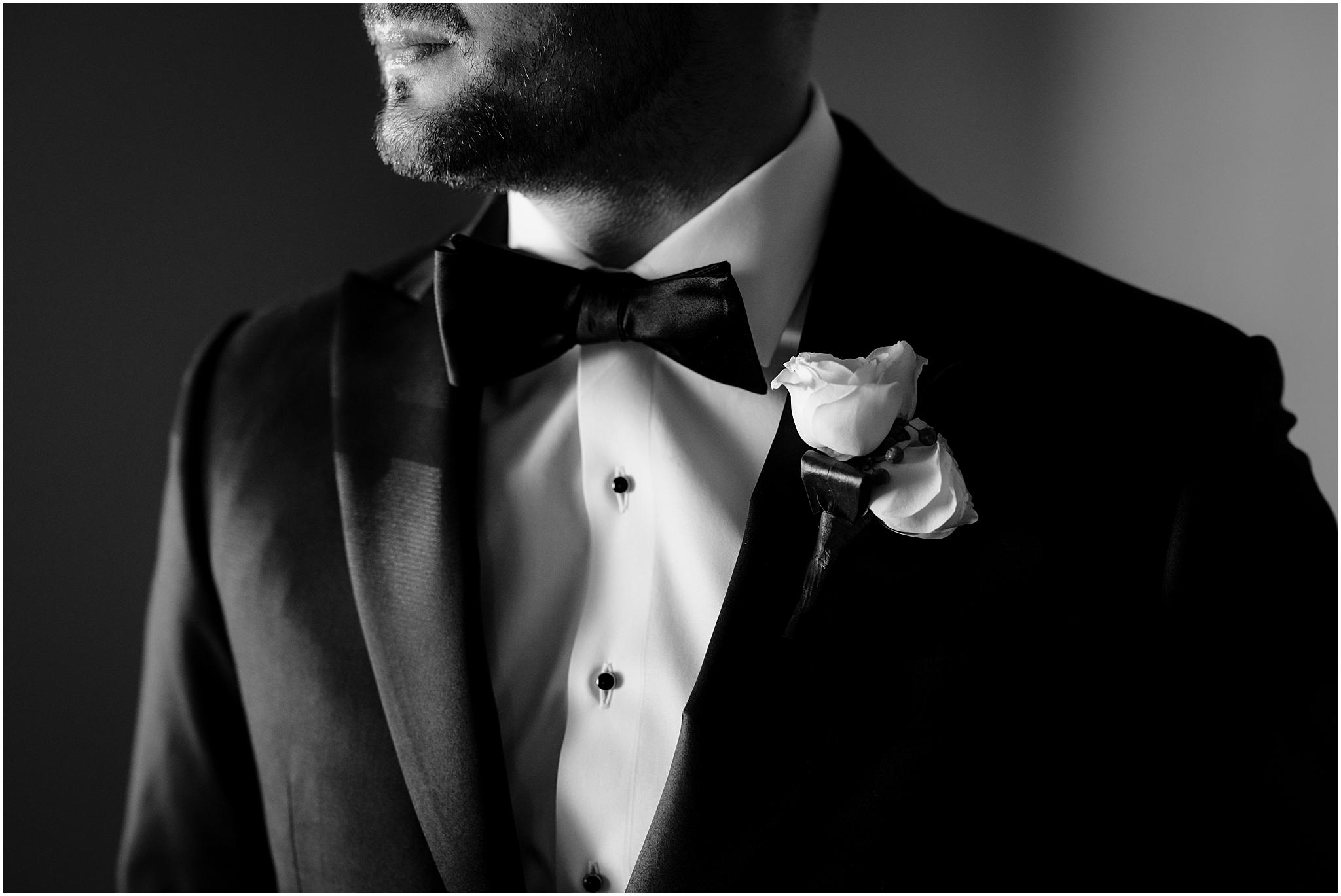 groom looks towards window with white rose boutonniere on black tux jacket