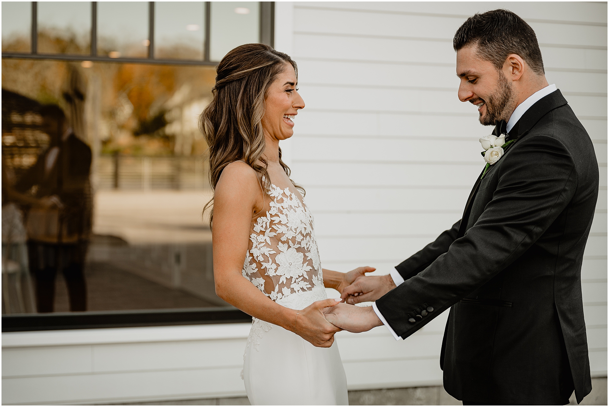 groom grins at bride looking at her wedding dress during first look on patio at the Waters Edge at Centerport