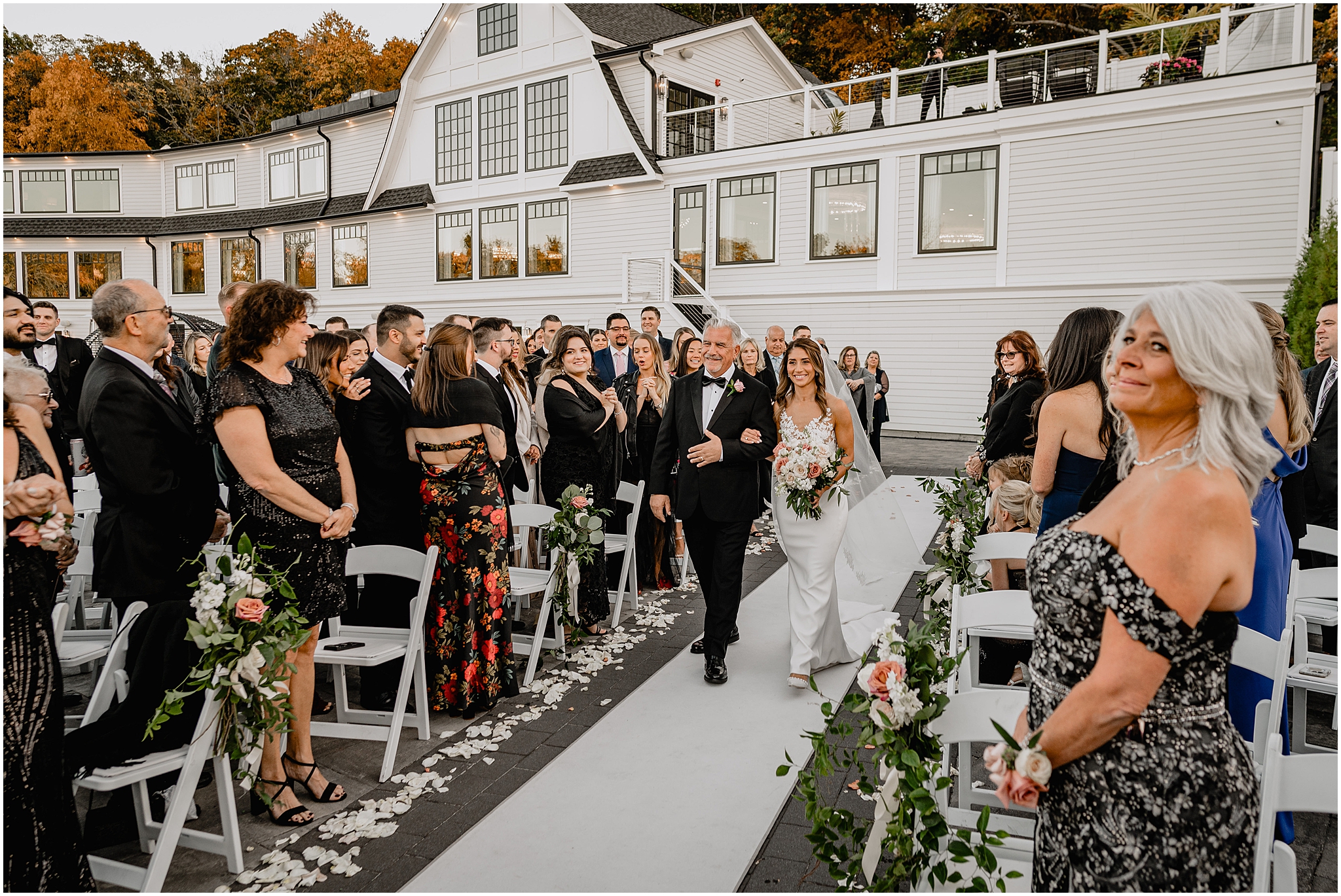 bride walks down aisle escorted by father for wedding ceremony on the patio of the Waters Edge at Centerport