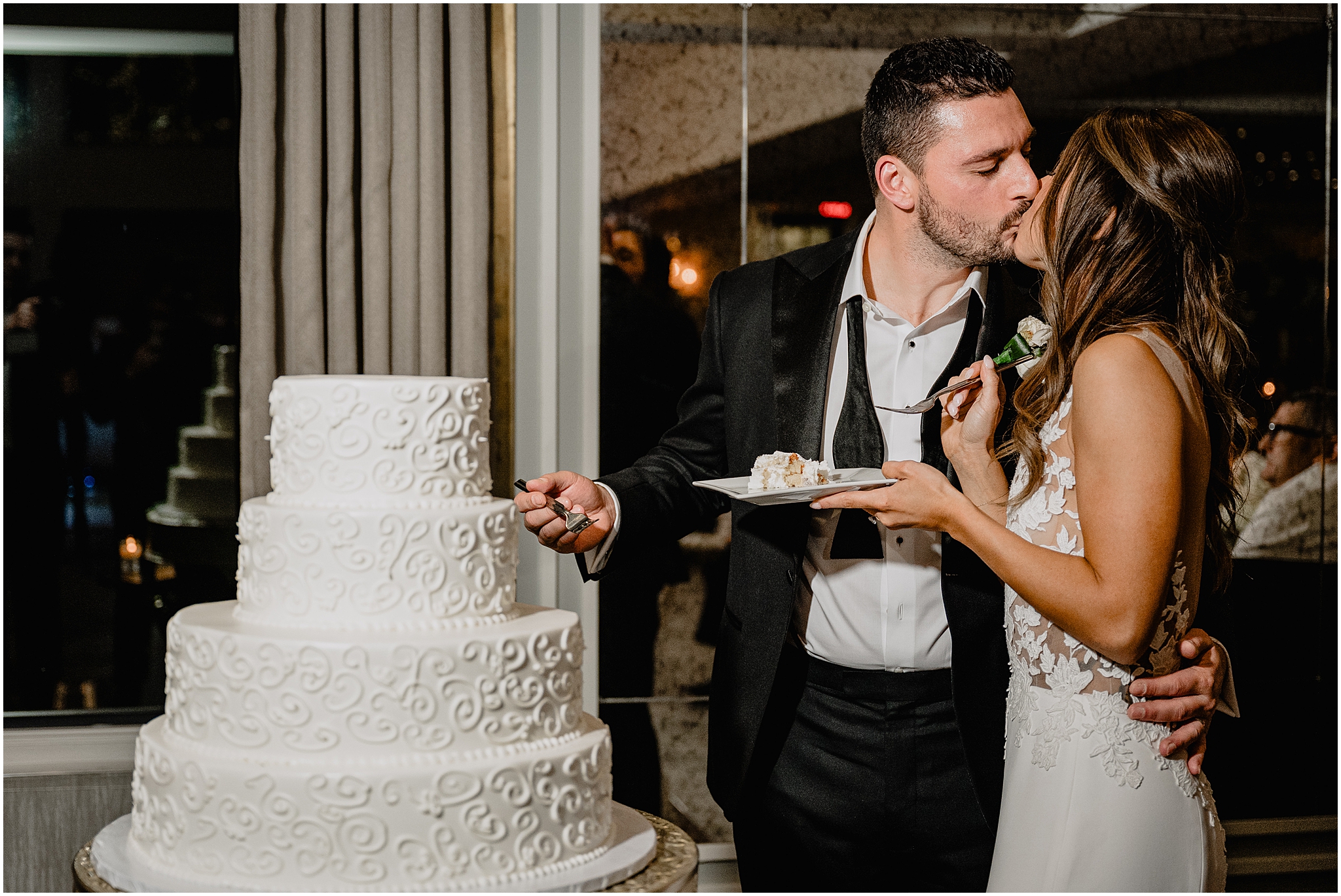  newlyweds kiss while cutting tiered wedding cake with white textured icing