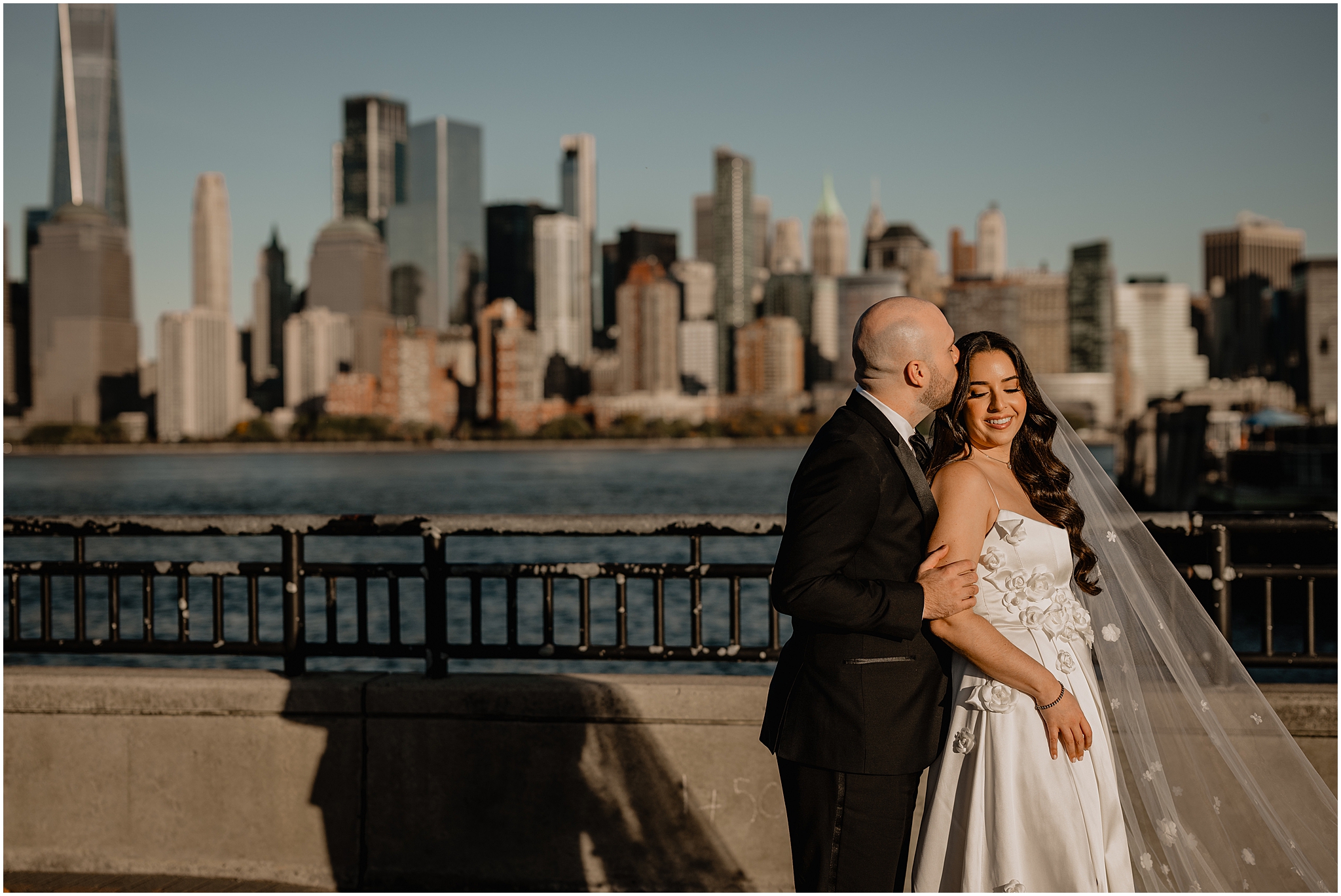 groom hugs bride from behind in front of Manhattan skyline in Jersey City
