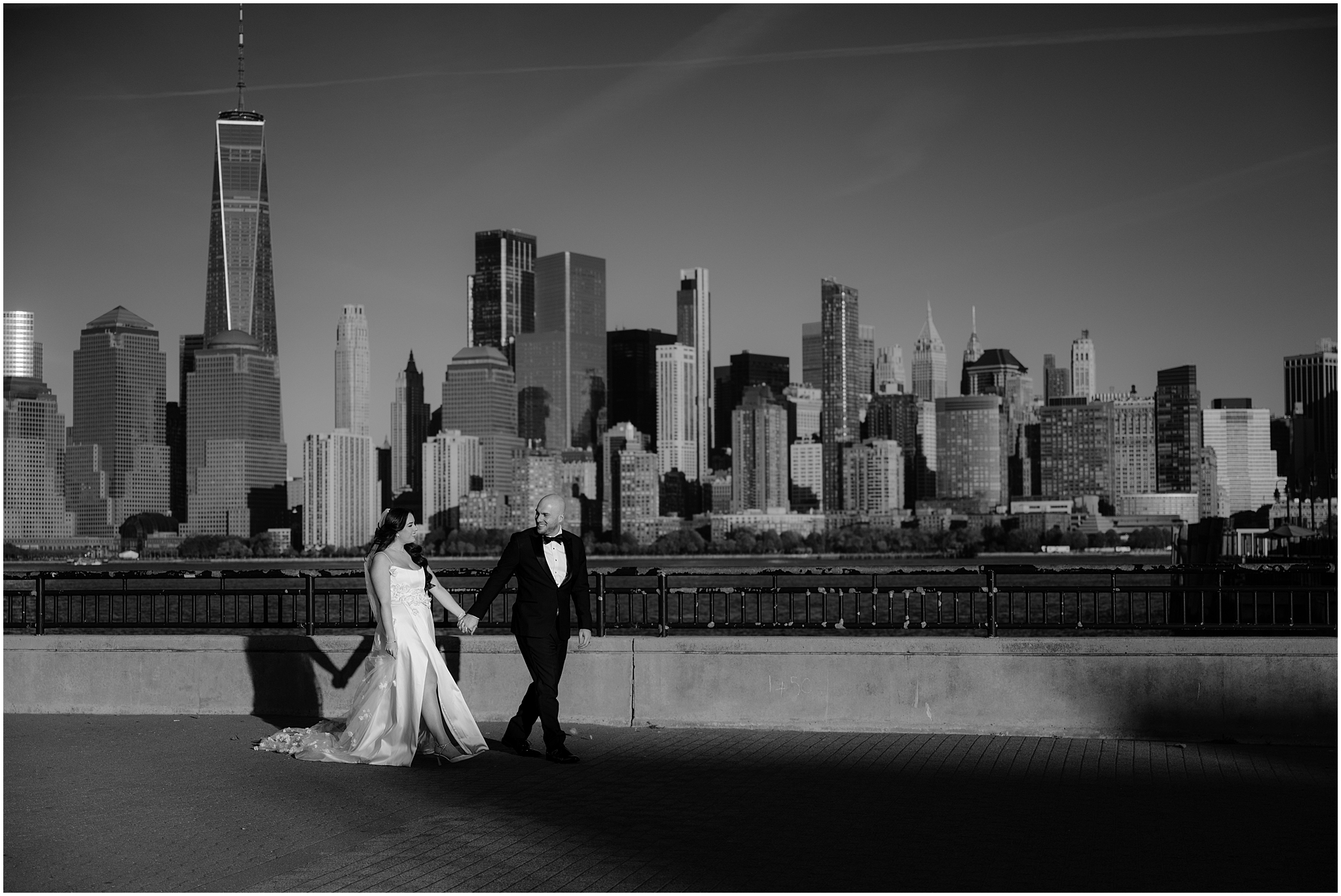 bride and groom hold hands walking along waterfront in front of Manhattan skyline 