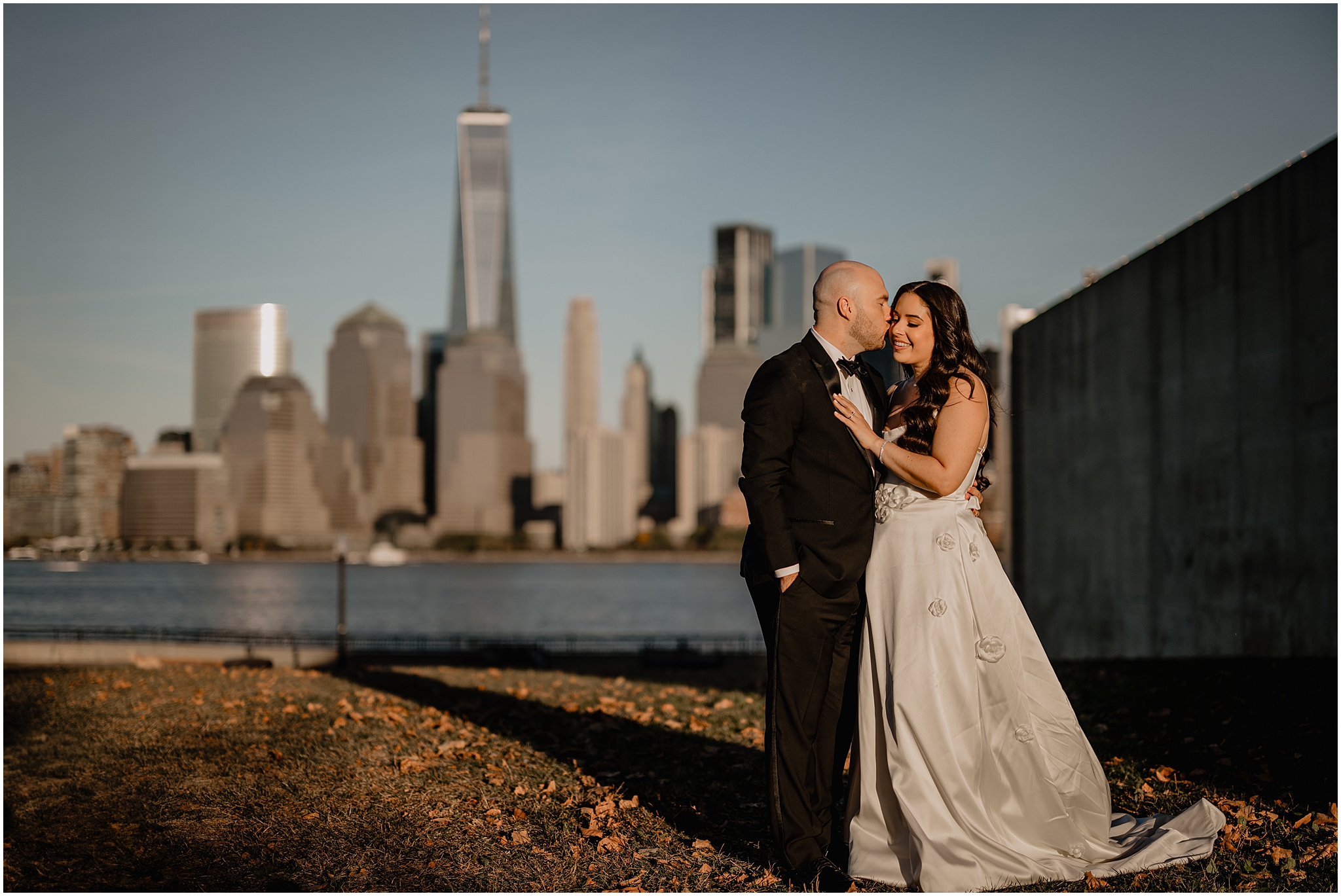 groom leans to kiss bride's cheek in front of NYC skyline in Jersey City