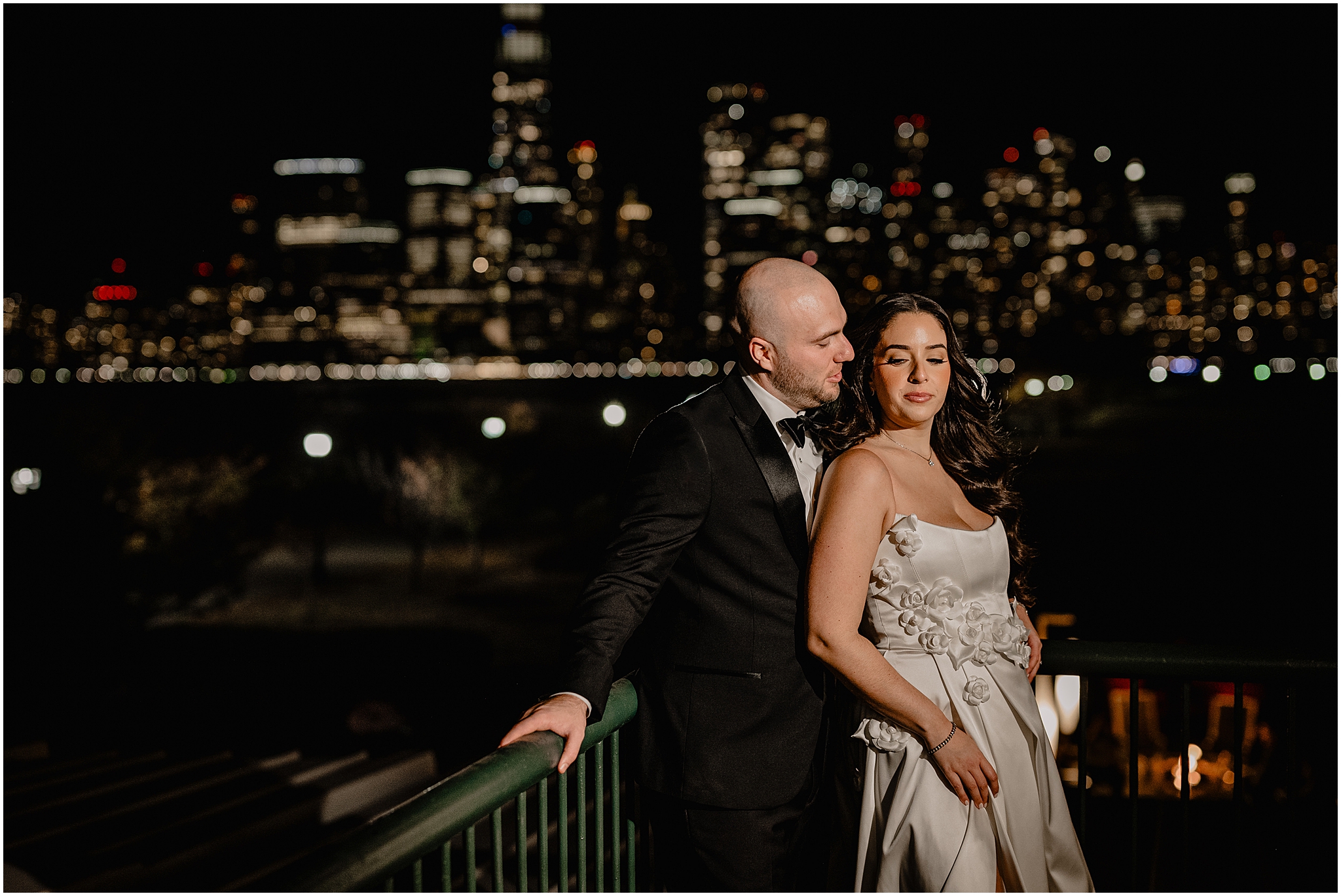 bride leans her back against groom in front of green railing at the Liberty House Restaurant at nighttime 