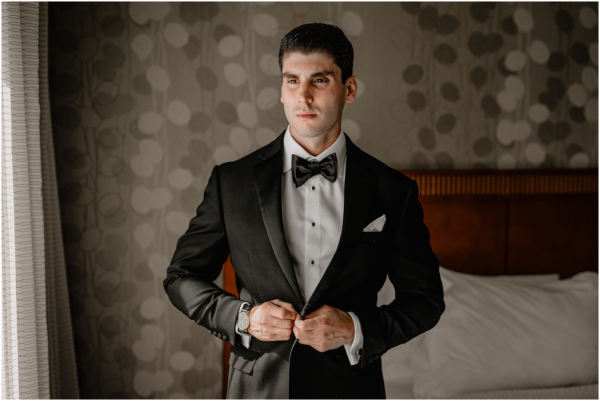 groom buttons up black suit jacket in front of bed in NJ hotel 