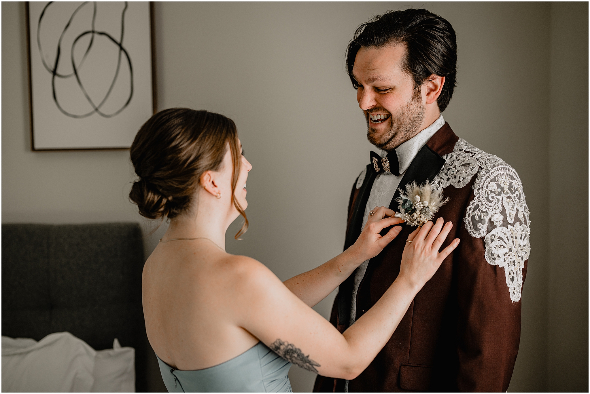 woman adjusts boutonniere for groom in burgundy suit jacket with lace accents 