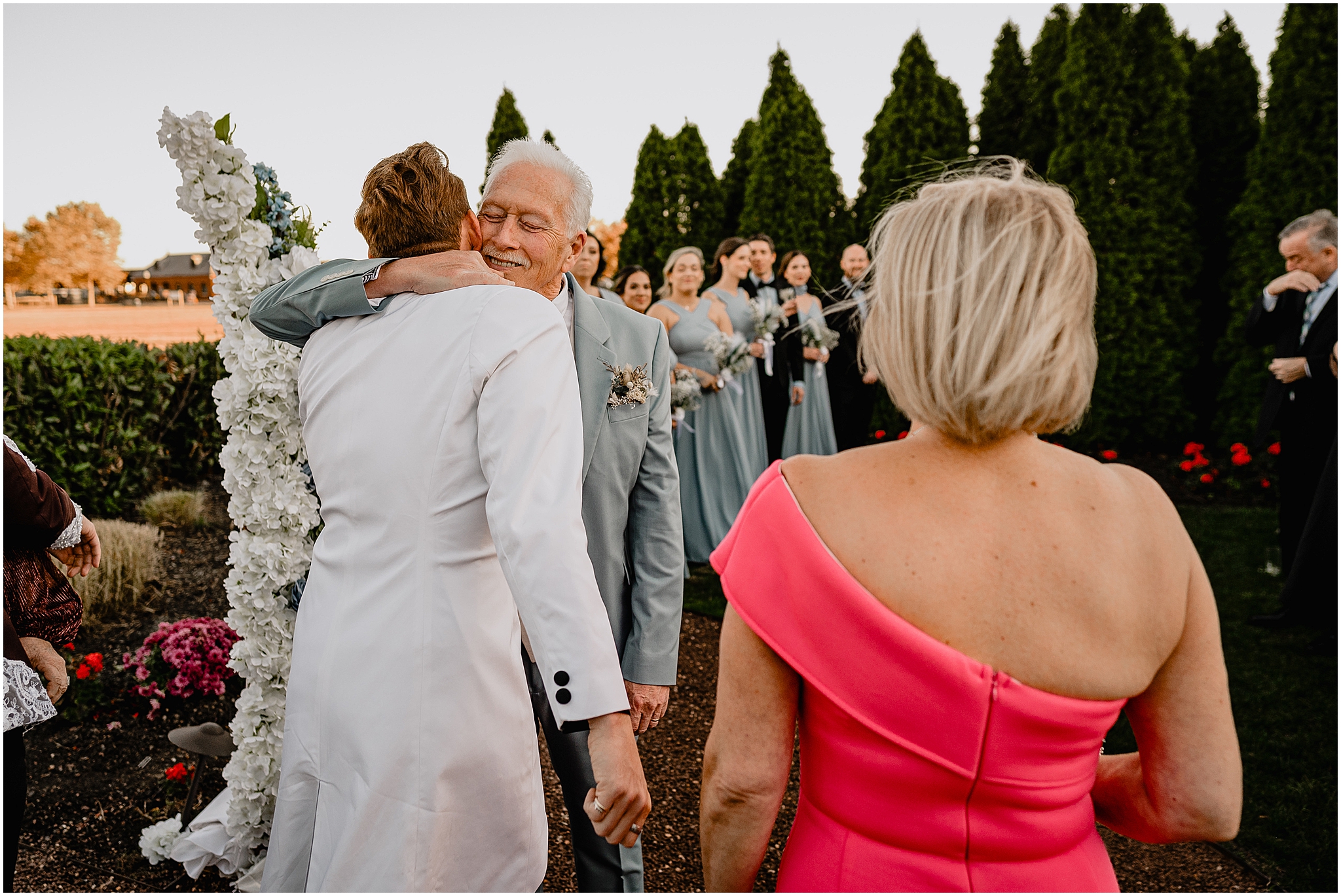 parents hug groom in white suit jacket in front of white coral arrangement at the Liberty House in Jersey City