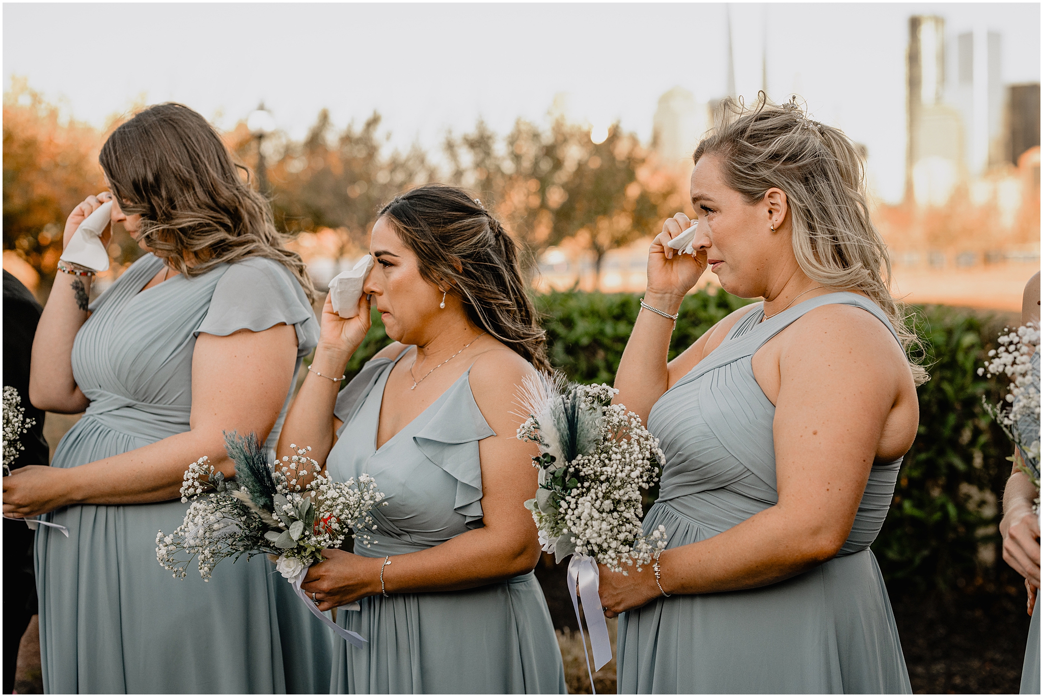 bridesmaids in dusty blue gowns wipe eyes with tissues during wedding ceremony 