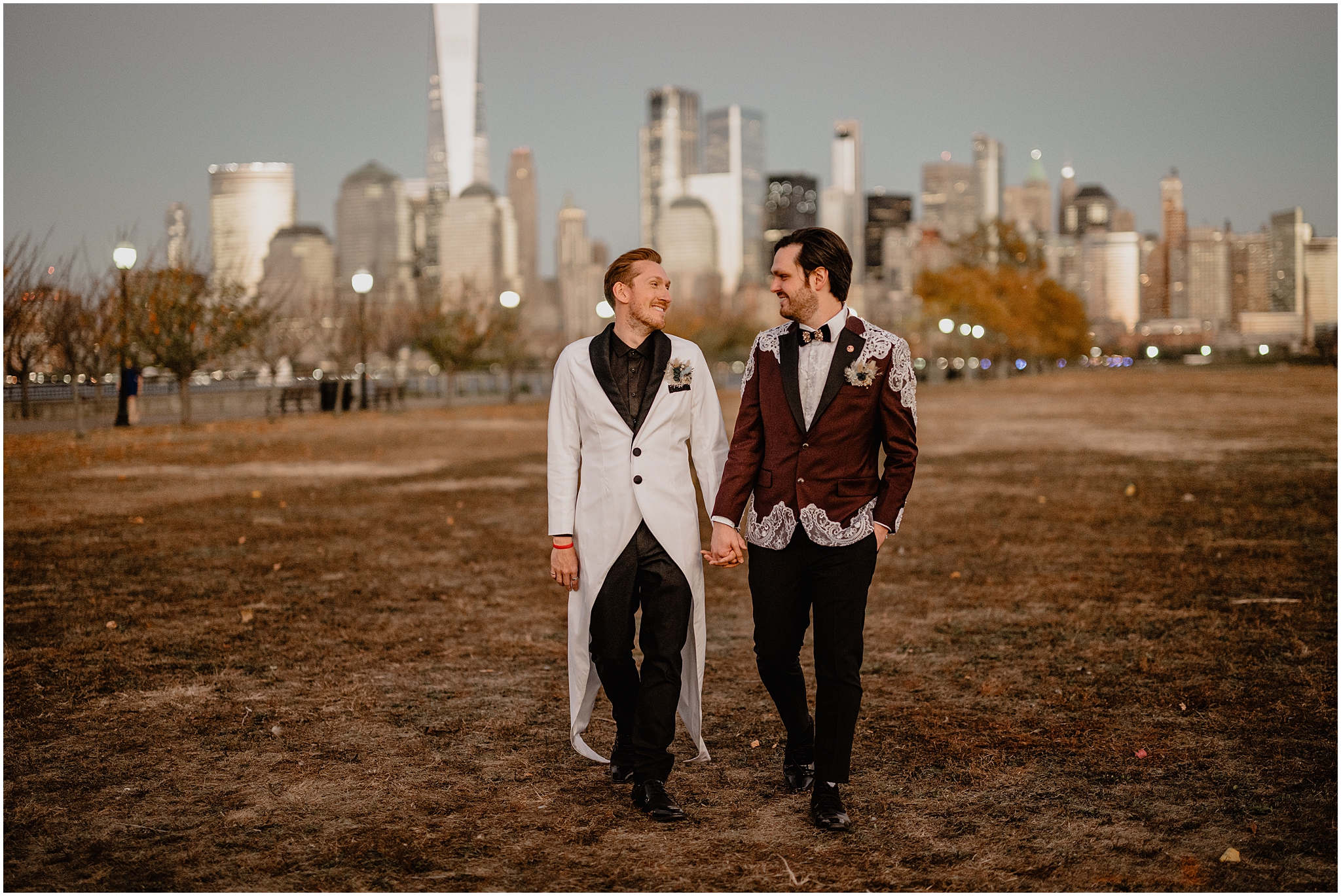 two men hold hands walking through park in Jersey City in front of NYC skyline