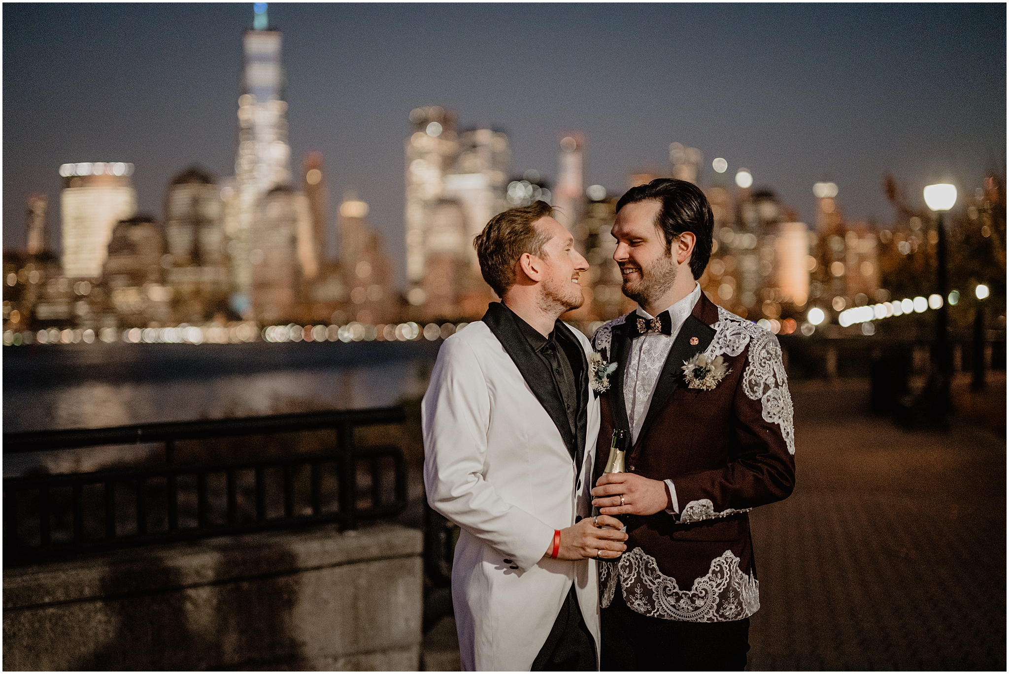 grooms smile at each other holding bottle of champagne in front of NYC skyline 