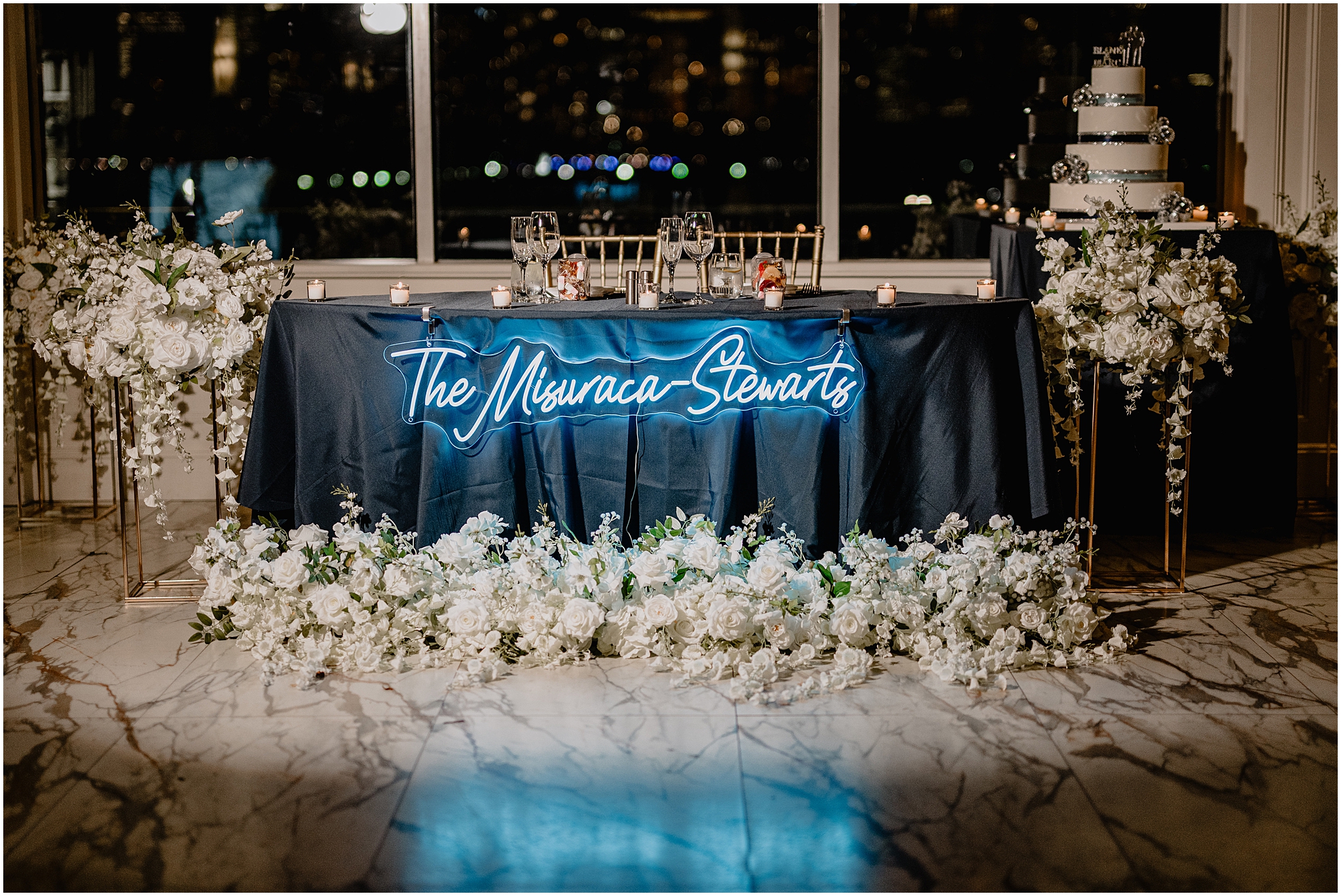 sweetheart table with white rose sprays in front of the table, custom neon sign and wedding cake behind them