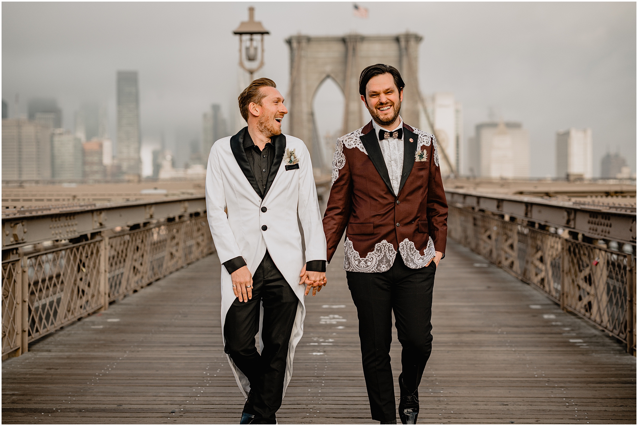 grooms hold hands laughing and walking on the Brooklyn Bridge during wedding portraits 