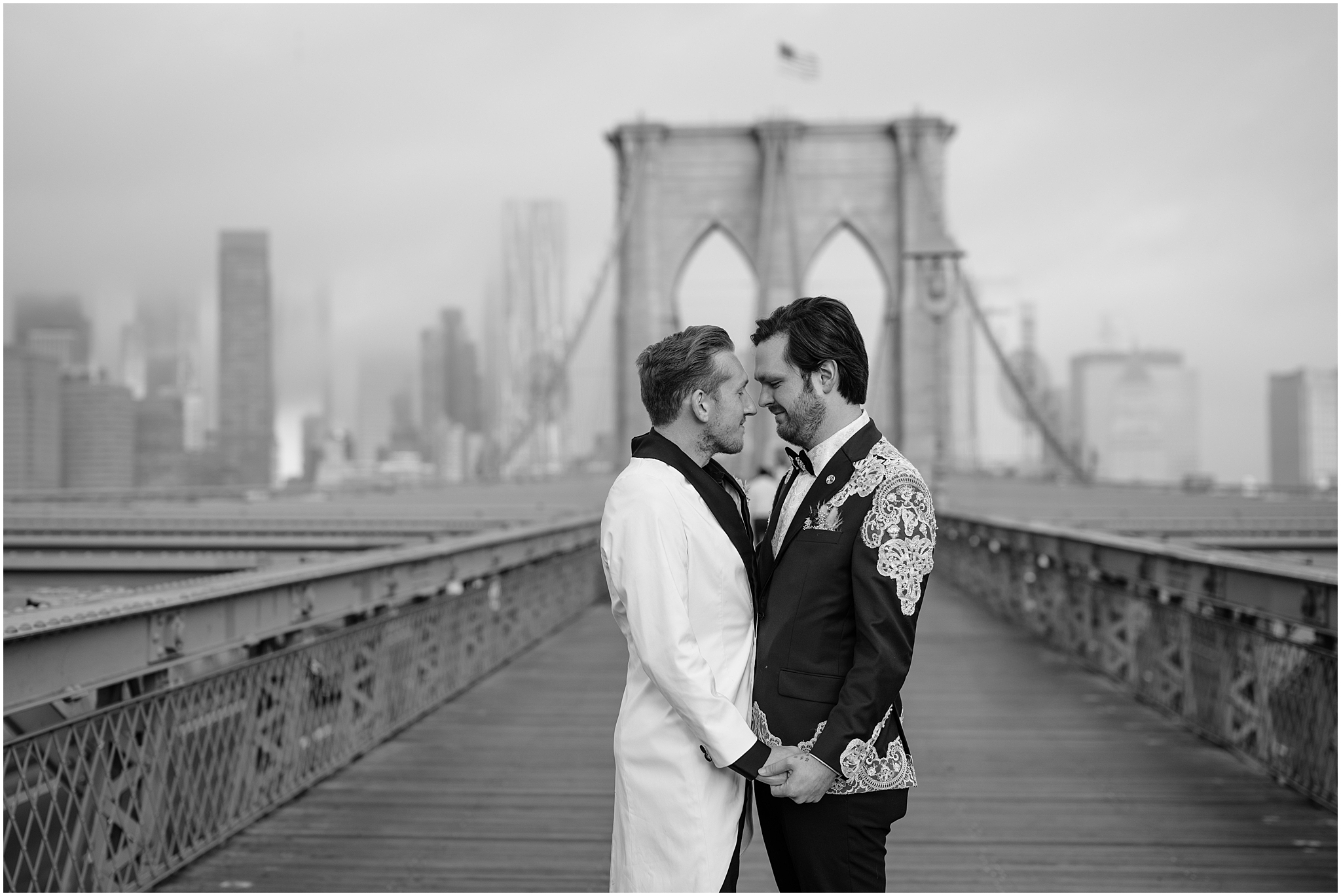 two grooms hold hands standing in front of Brooklyn Bridge during wedding portraits 