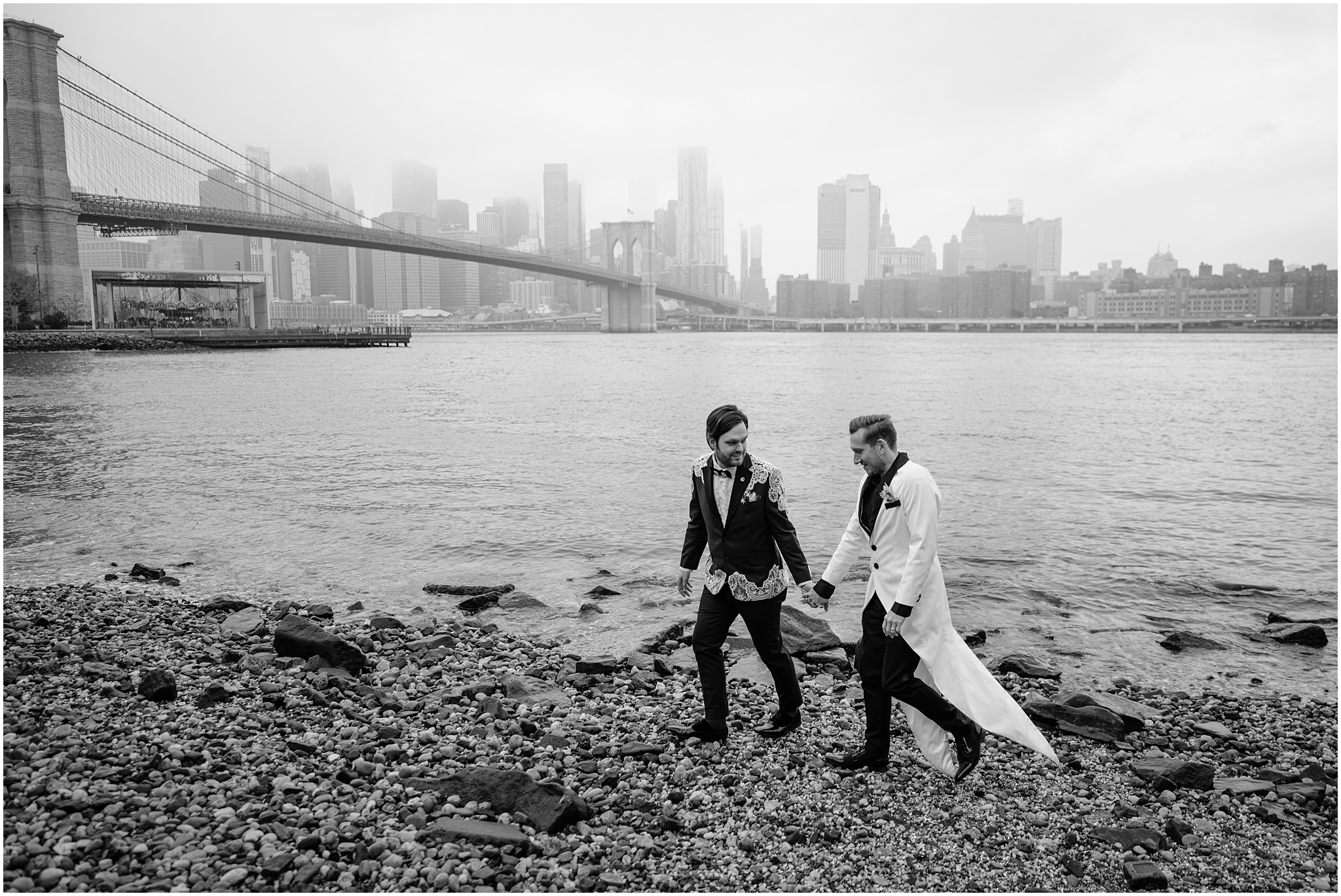 two grooms hold hands walking on pebbles in front of the Brooklyn Bridge amongst NYC waterfront 