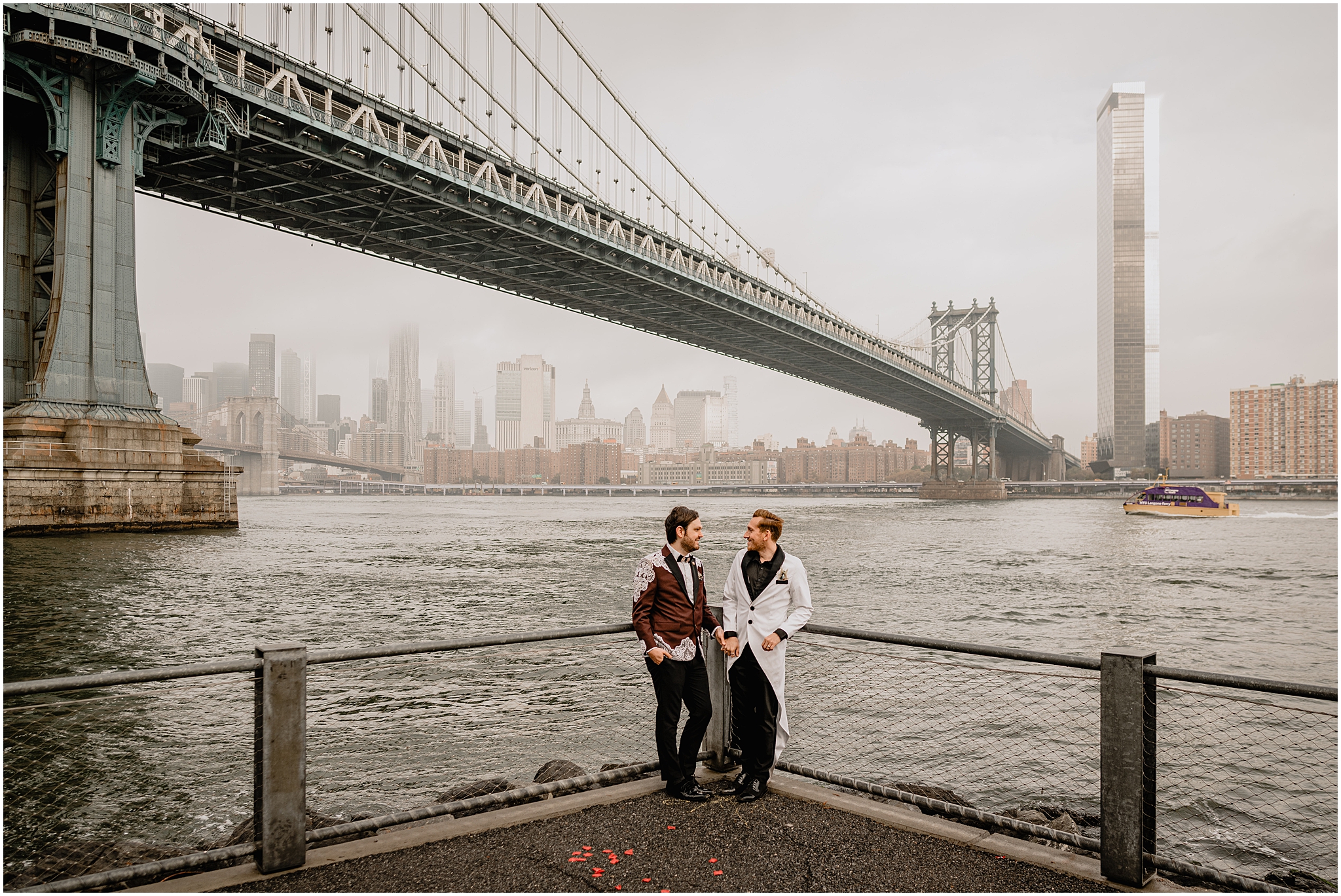 grooms lean against corner of fence line in front of the Brooklyn Bridge 
