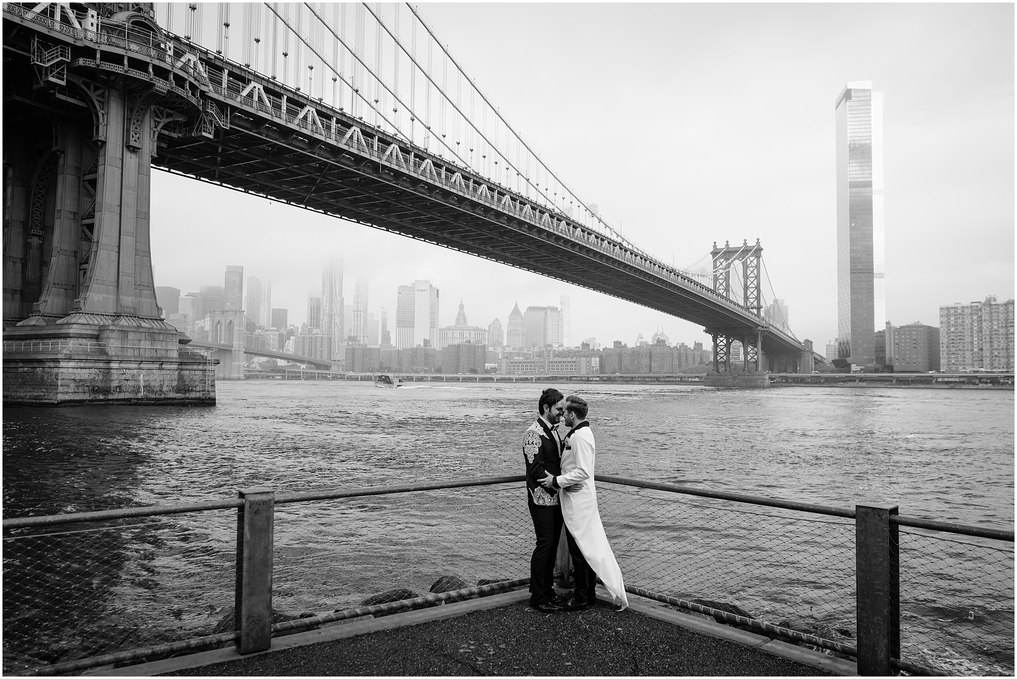two grooms hug in corner of overlook in front of the Brooklyn Bridge on wedding day