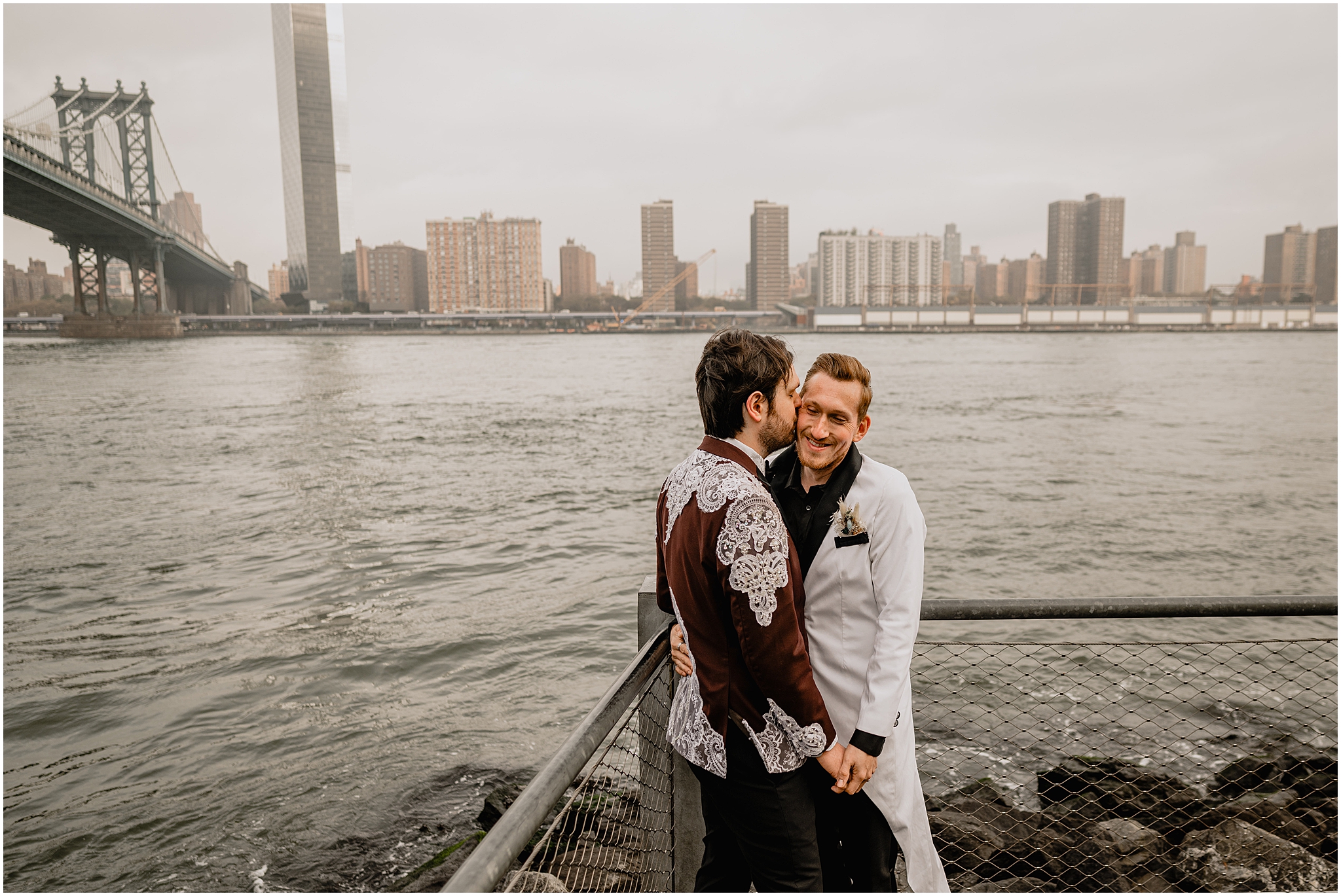 two grooms hug in corner of fence in front of Brooklyn Bridge 