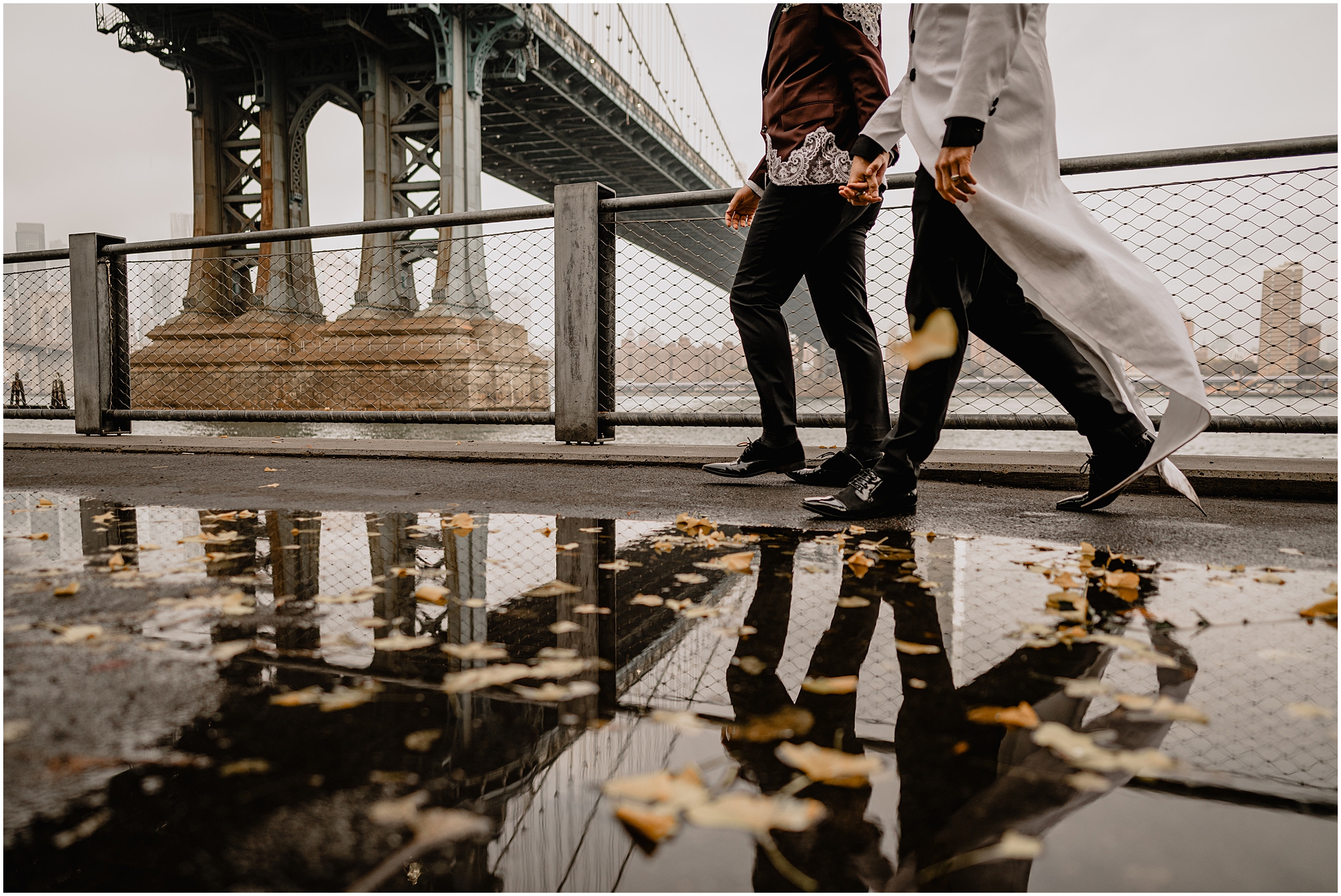 two grooms hold hands walking under bridge reflected in the water 