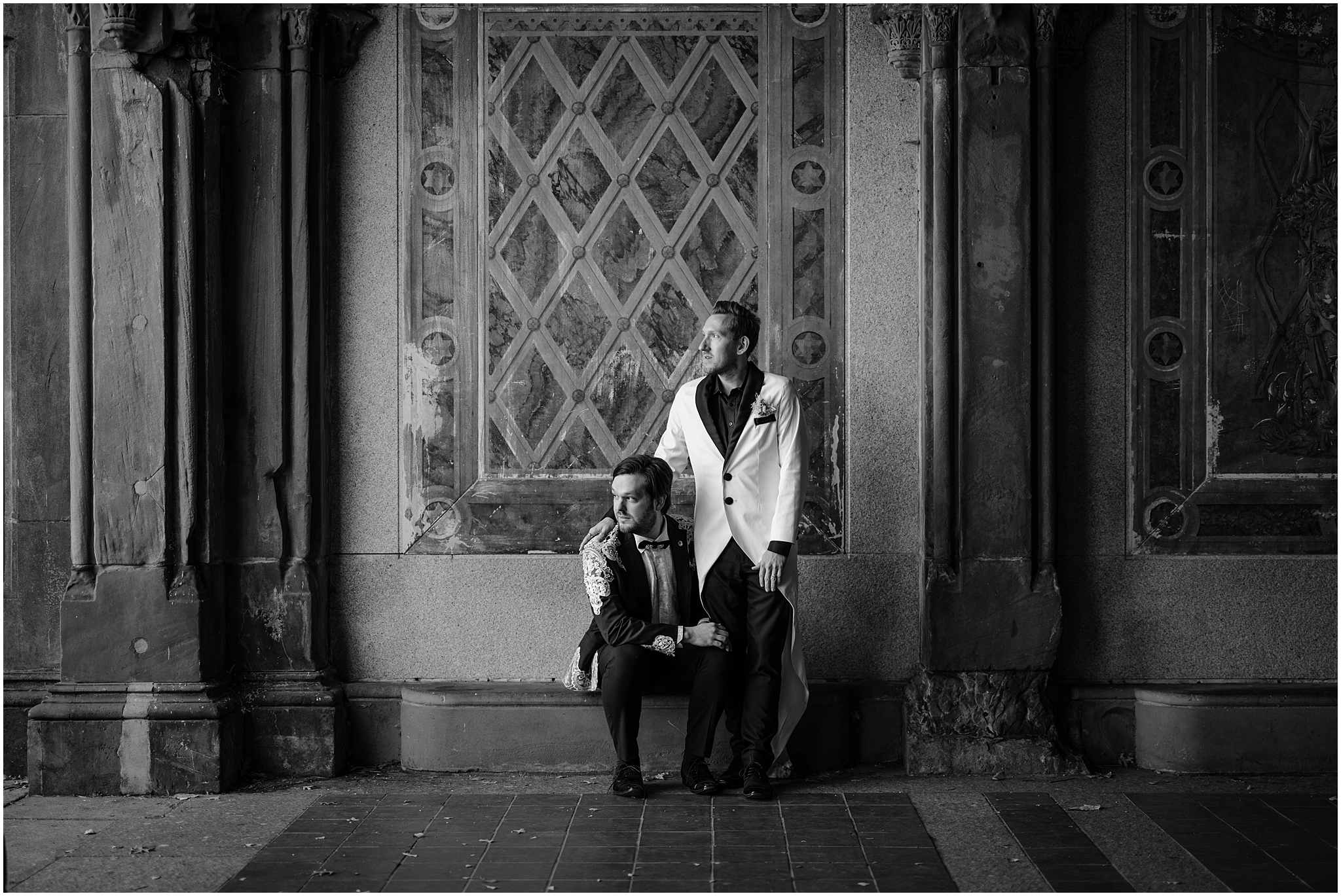 groom in white jacket stands with groom in burgundy jacket holding his leg standing in front of mural in Bethesda Terrace 