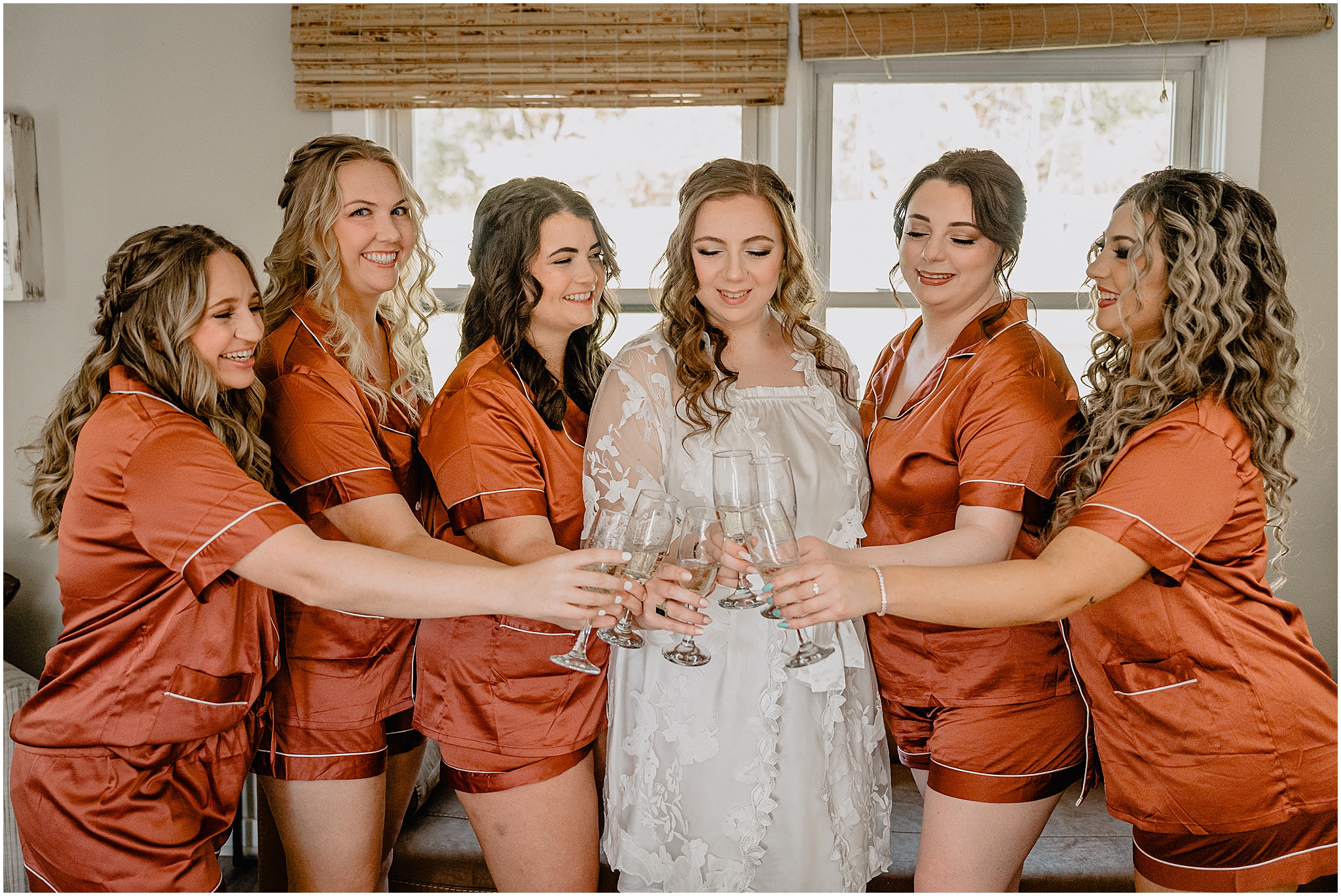 bridesmaids in orange silk pajamas smile at bride while toasting champagne glasses 