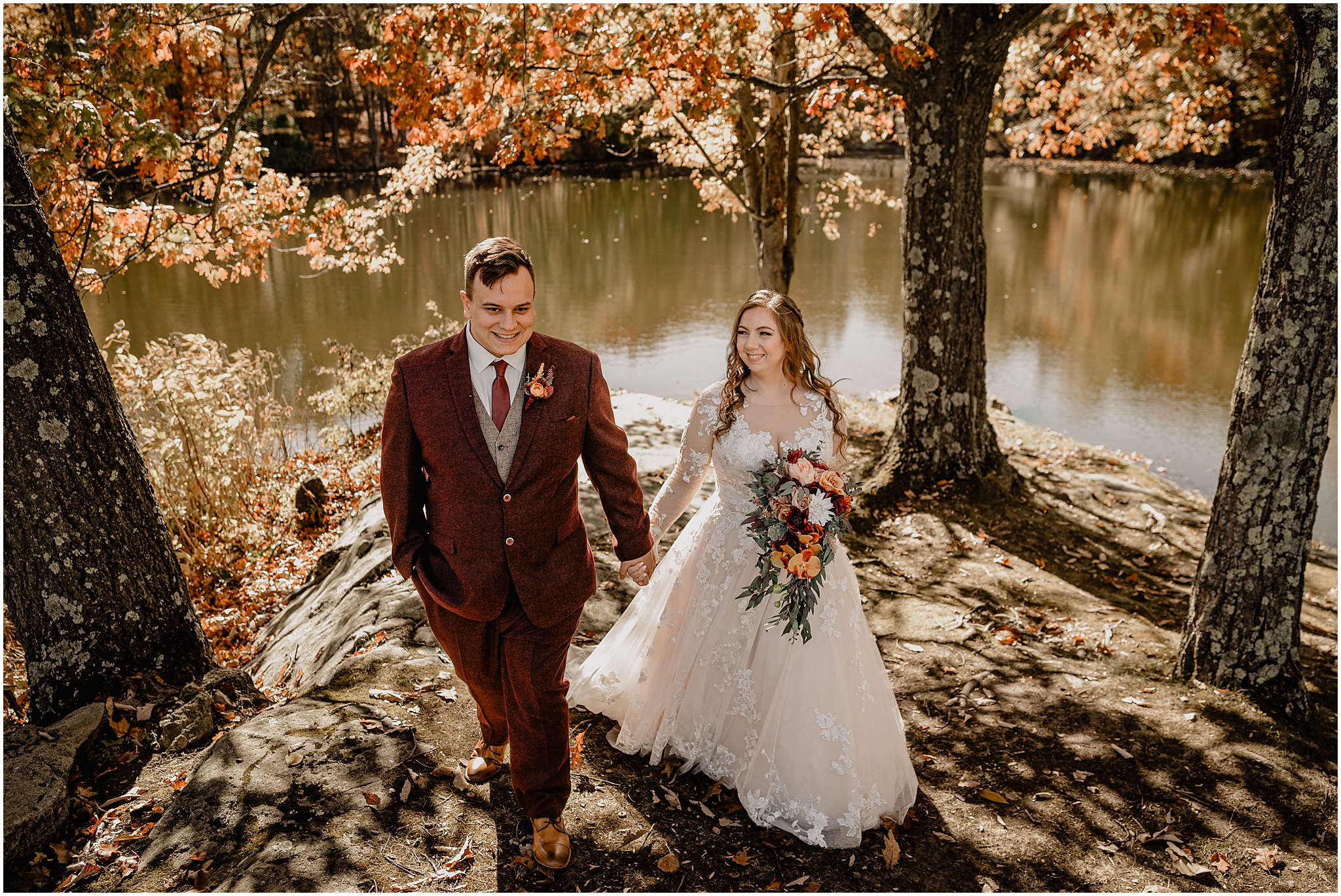 groom in burgundy suit holds hands walking with bride on rocks outside the Barn at Perona Farms