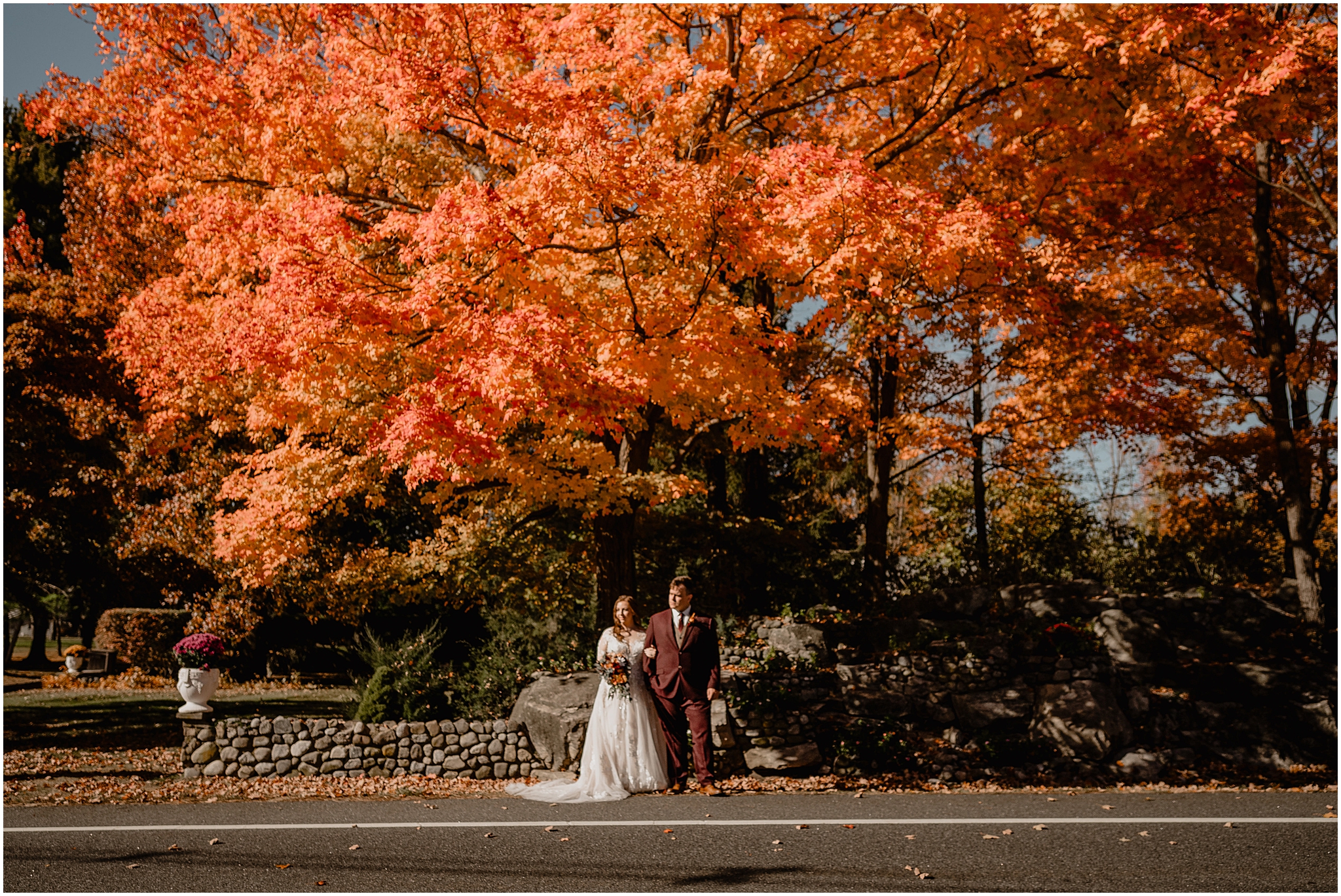 newlyweds stand holding hands under orange leaves on trees at the Barn at Perona Farms