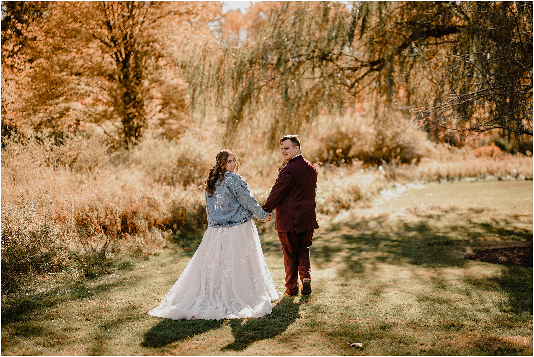 bride in jean jacket holds hands with groom in burgundy suit walking under willow tree in New Jersey
