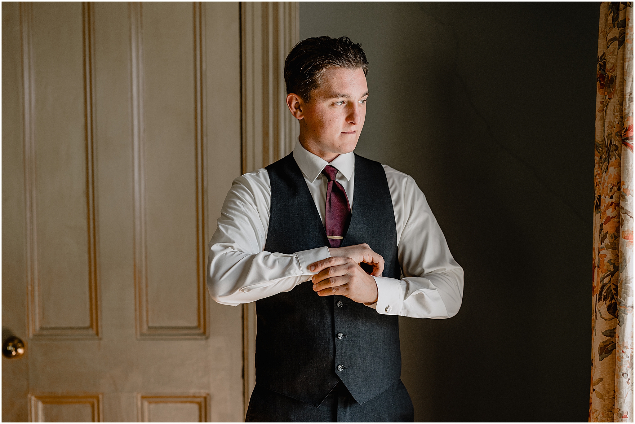 groom adjusts cufflinks in dark grey suit with burgundy tie looking out window 